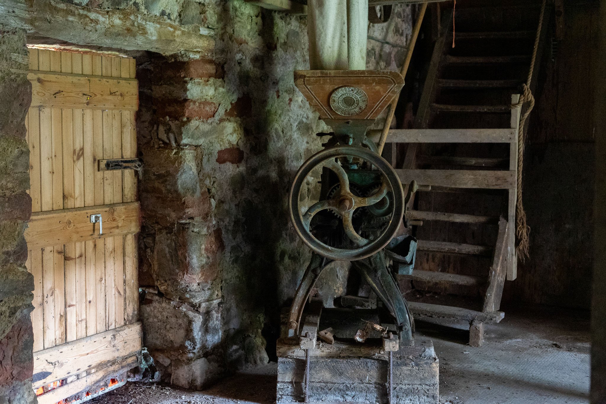 An old vintage mechanical grape crusher or press machine inside a rustic, stone-walled cellar with wooden stairs in the background.