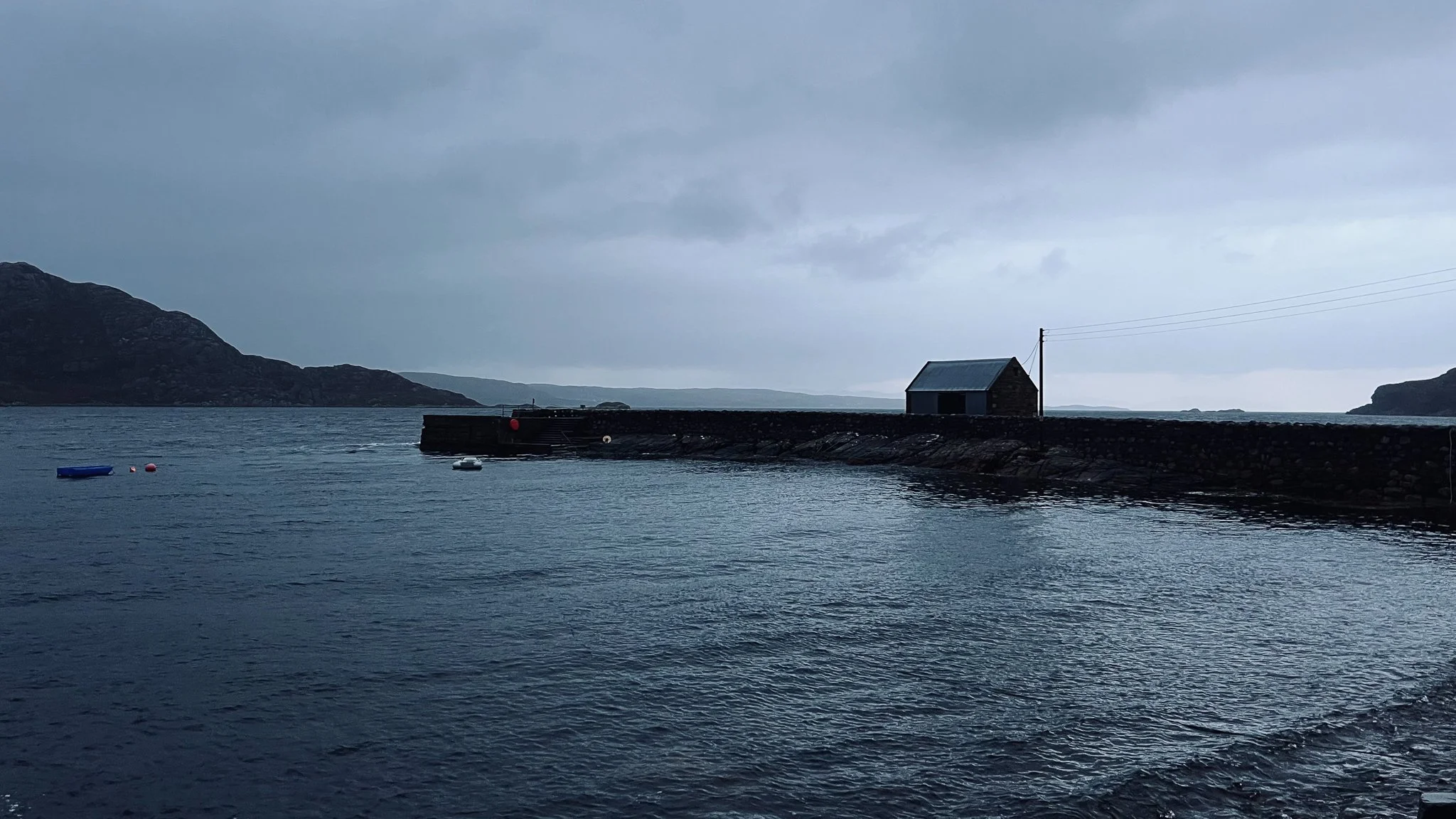 A coastal scene with a stone pier extending into the water, a small wooden building on the pier, and hills in the background. The sky is overcast, and there are a few small boats floating near the pier.