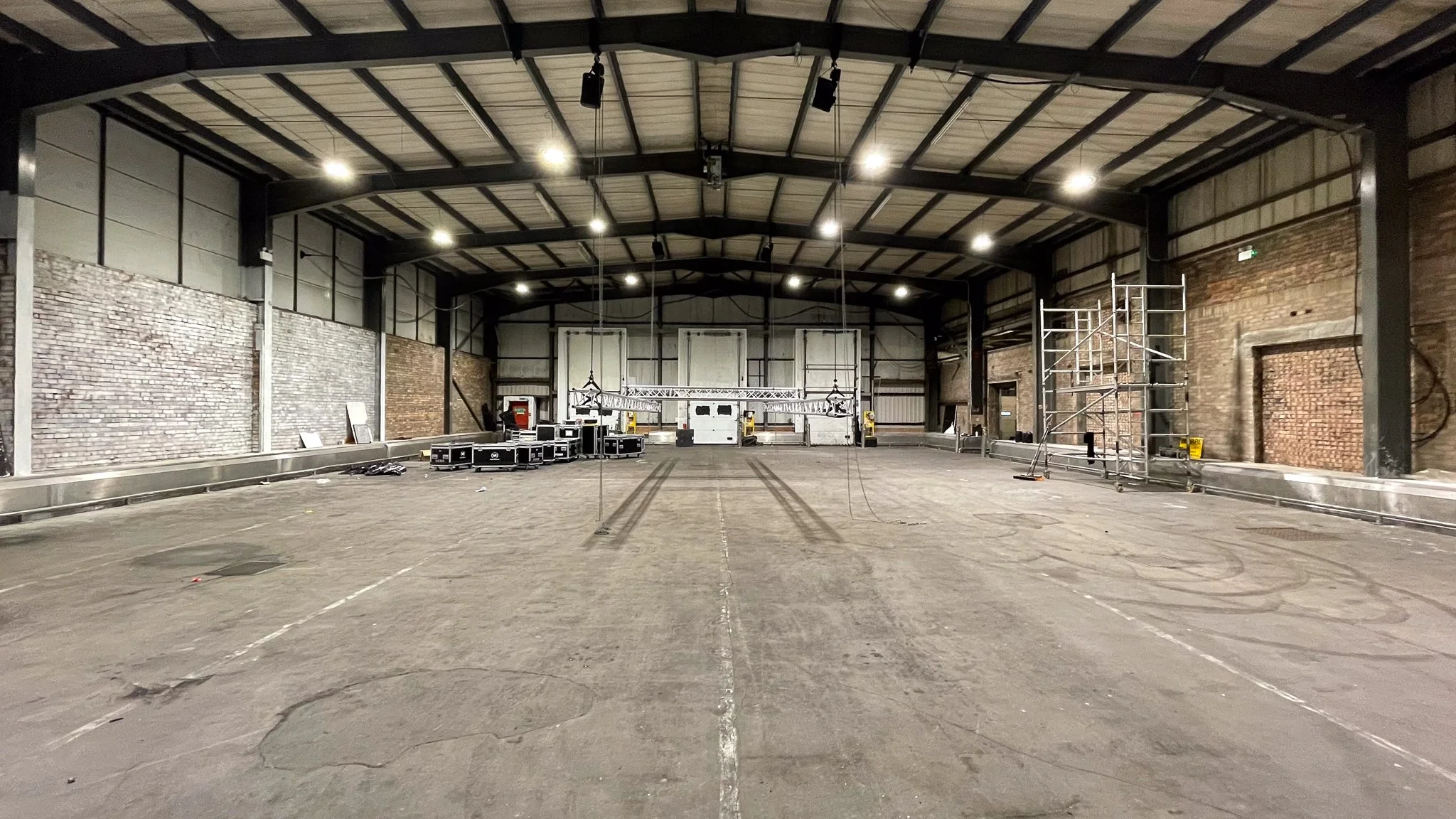 An empty industrial event space or warehouse with high ceilings, exposed brick walls, and scaffolding on the right side. Temporary lighting and equipment are set up, with a large metal truss hanging from the ceiling, likely for lighting or sound equi