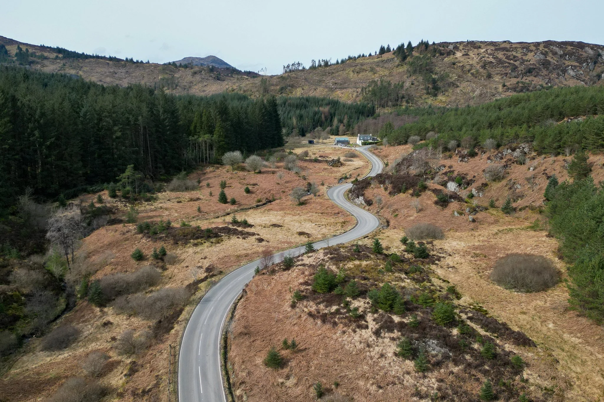 A winding mountain road through a rural landscape with trees, shrubs, and hills in the background.