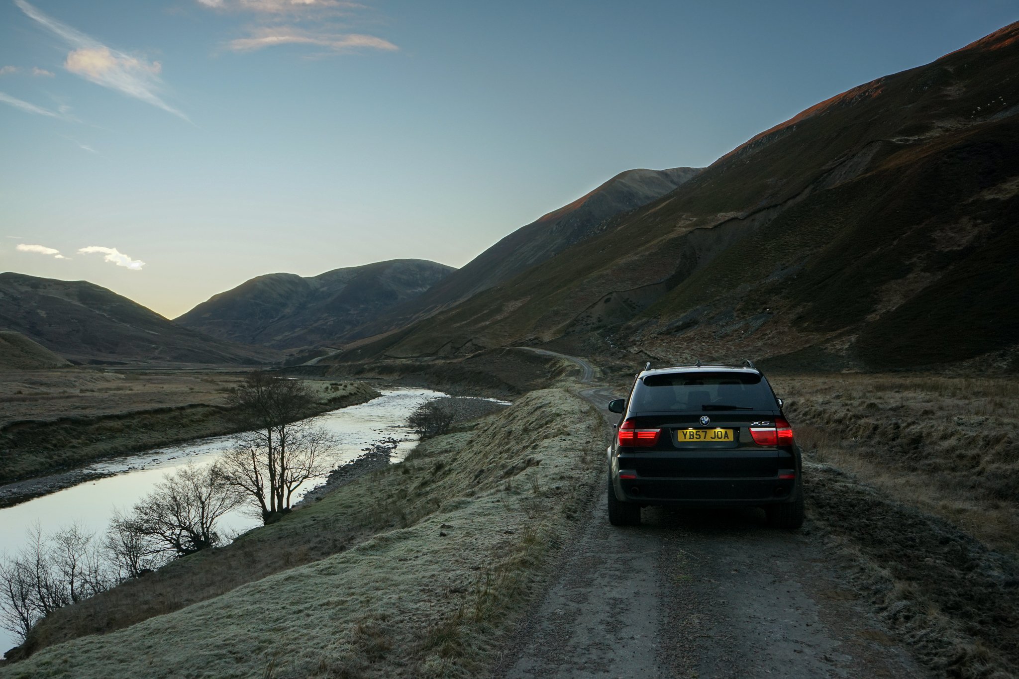 A black BMW X5 parked on a narrow dirt road beside a river in a mountainous landscape at dusk.