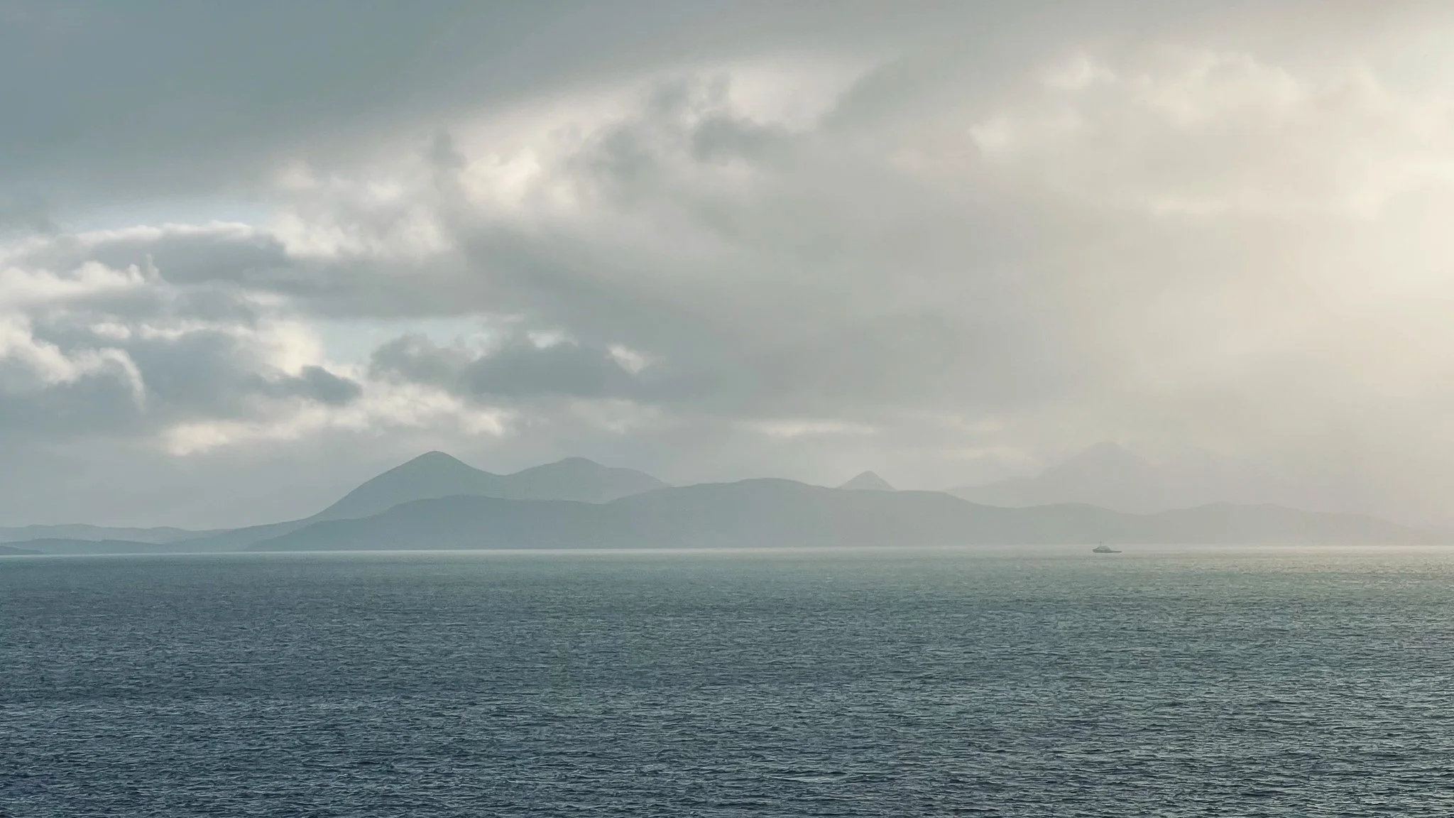 Ocean with mountains in the distance under cloudy sky, sunlight breaking through clouds, a small boat visible on the water.