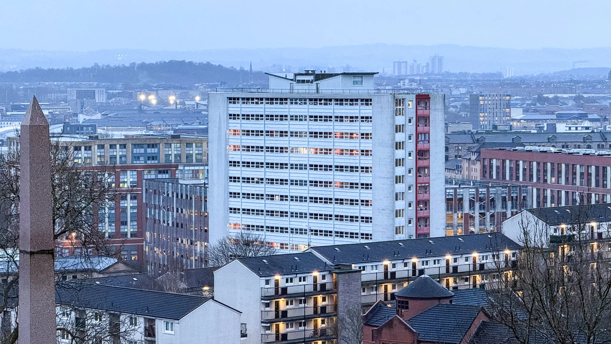 Cityscape with multiple high-rise buildings, some with illuminated windows, at dusk or early evening.
