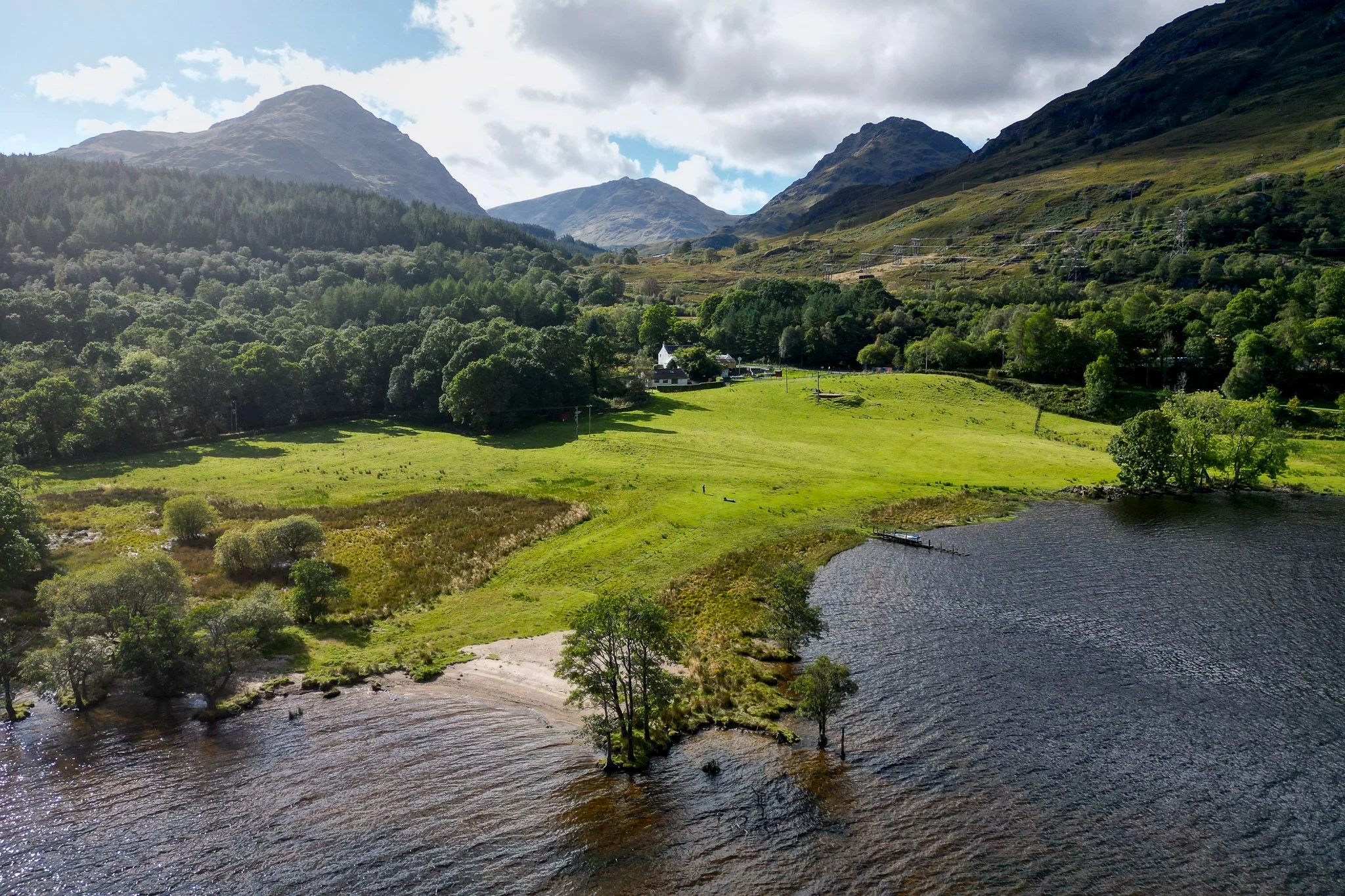 A scenic view of a lake with small trees along the shoreline, green grass fields, and mountains in the background under a partly cloudy sky.