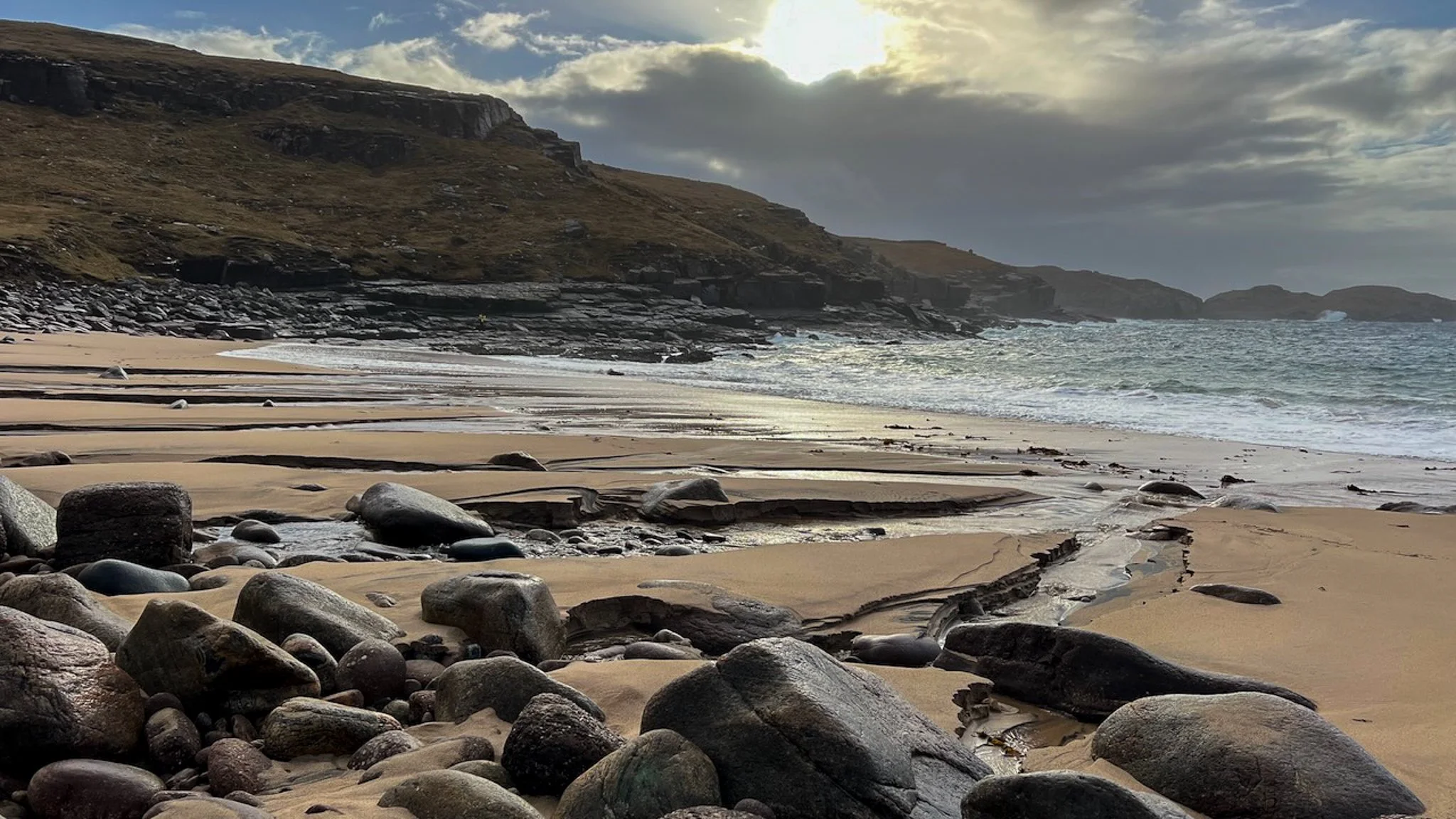 A sandy beach with large rocks in the foreground, gentle waves, and a cloudy sky with the sun partially visible behind clouds.