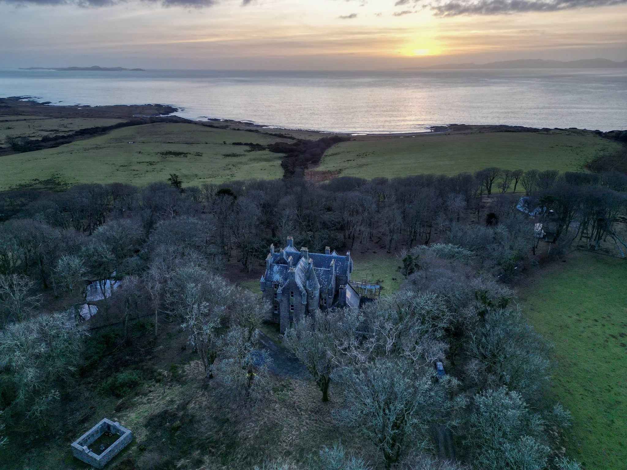 An aerial view of a large, old mansion surrounded by trees, with green fields and the ocean in the background during sunset.