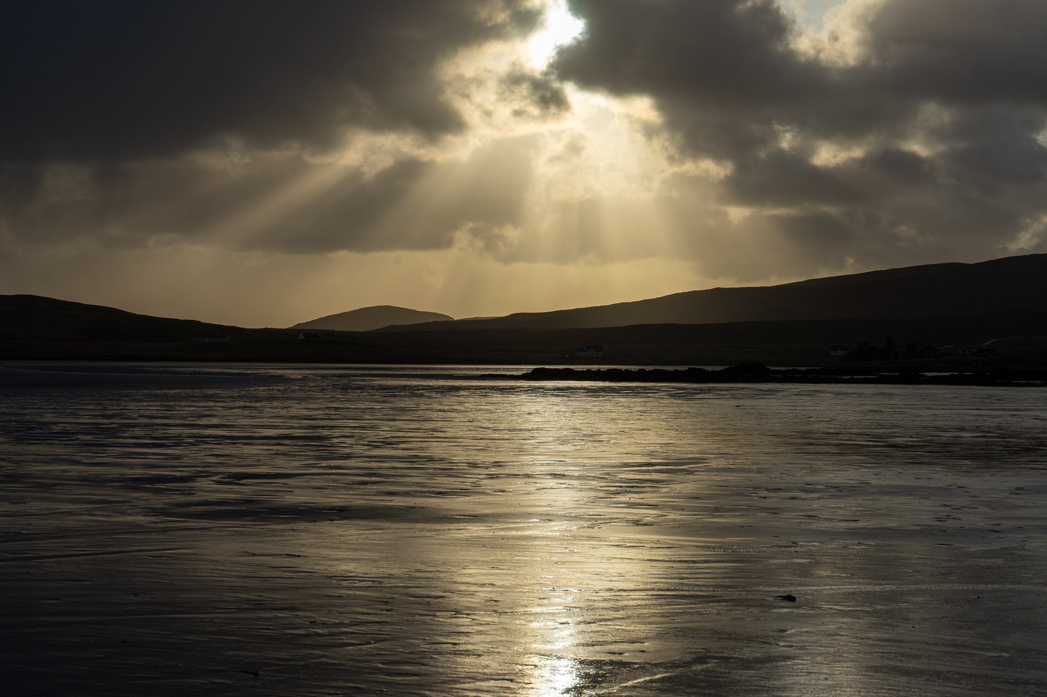 Sun rays breaking through dark clouds over a calm body of water with hills in the background.