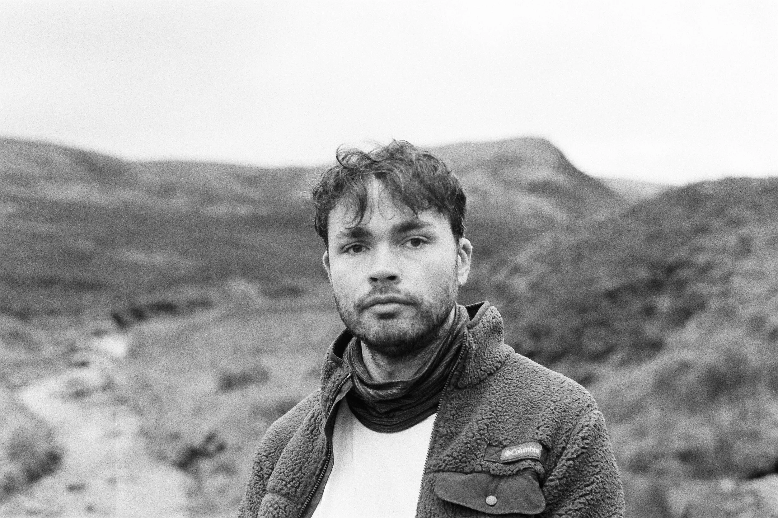 Black and white photo of a man with short hair and a beard, standing outdoors in a hilly landscape.