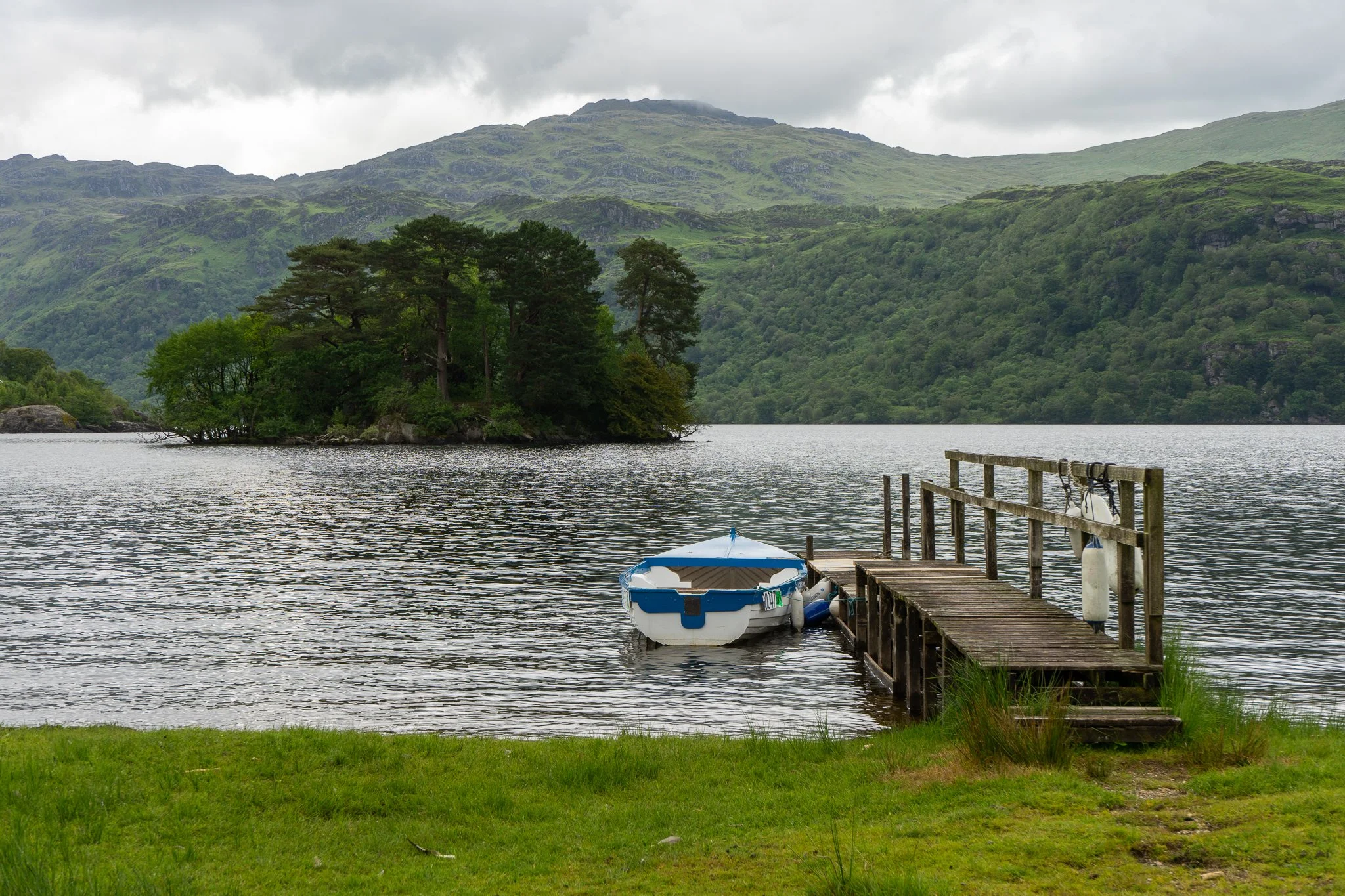 A peaceful lakeside scene with a small wooden dock and a boat attached to it. In the background, there is a tree-covered island and rolling green hills under a cloudy sky.