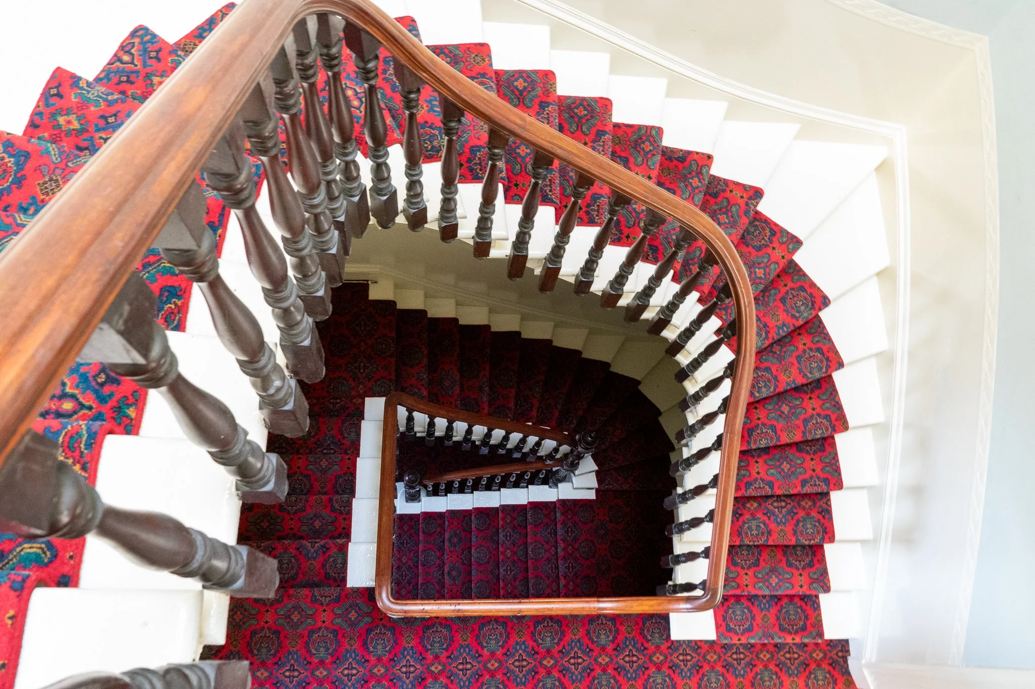 Top-down view of a spiral staircase with a red patterned carpet, black spindles, and a wooden handrail.