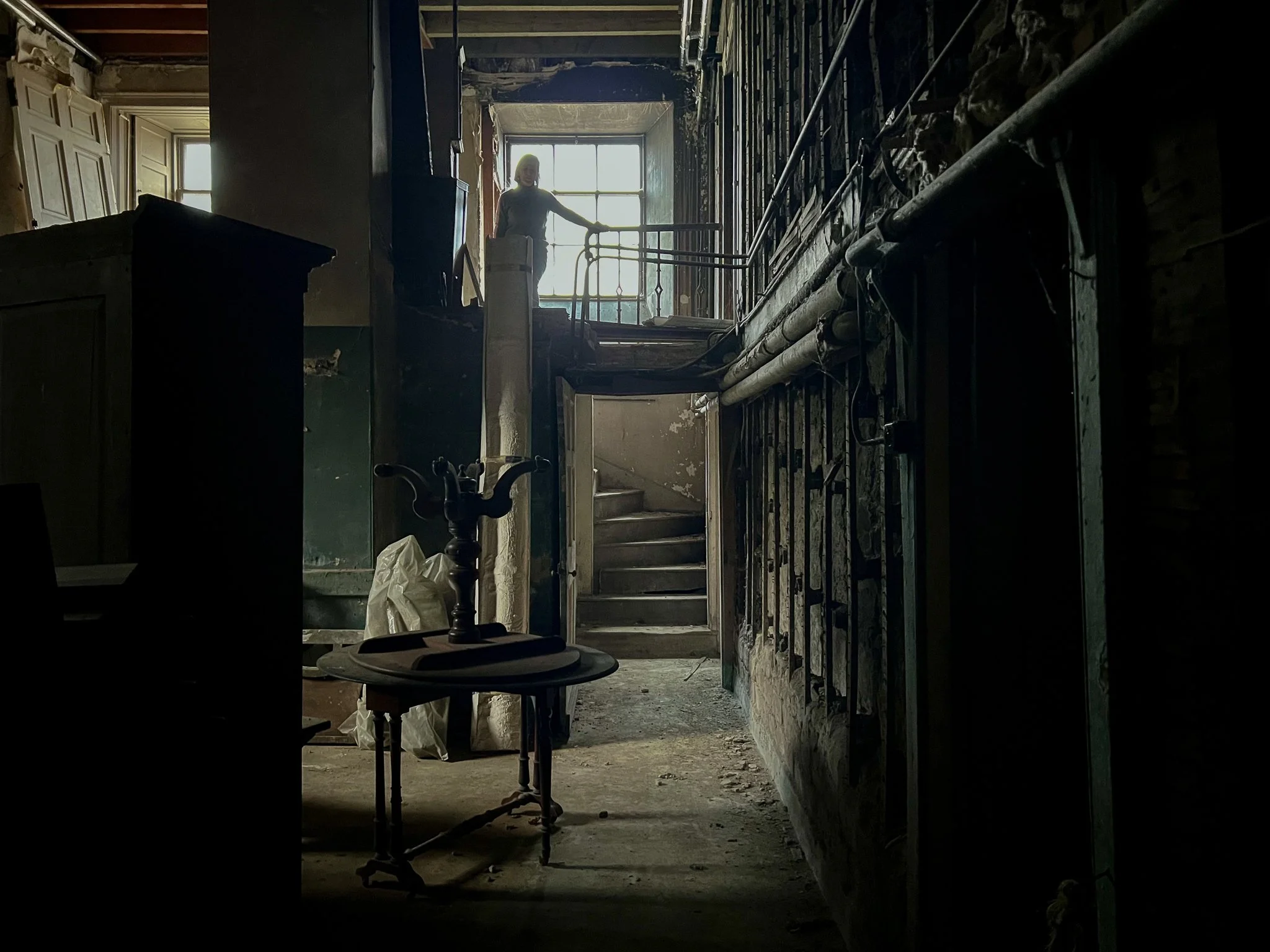 Dark and worn interior of a building under renovation, with exposed pipes on the wall, a staircase in the background, a window at the top of the stairs, and a person standing at the window.