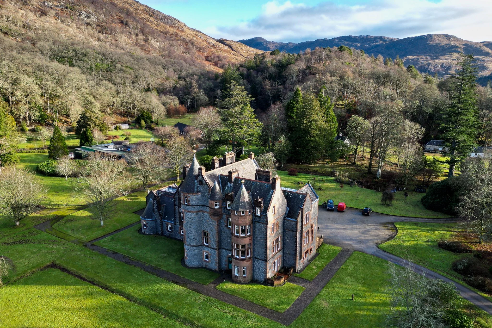 A large castle-like building with multiple turrets, set in a lush, green landscape surrounded by trees and mountains in the background.