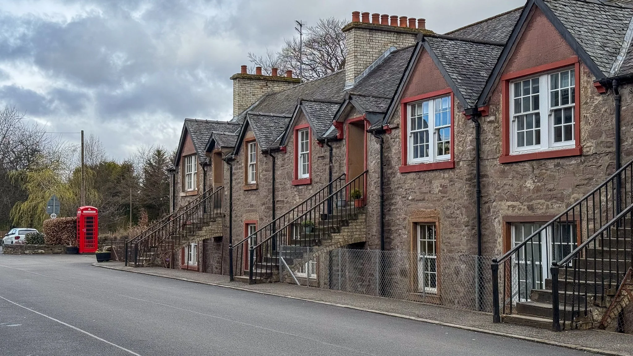 A row of stone houses with staircases leading up to the front doors, some with potted plants, and a traditional red telephone booth on the sidewalk. There are leafless trees and cloudy sky in the background.