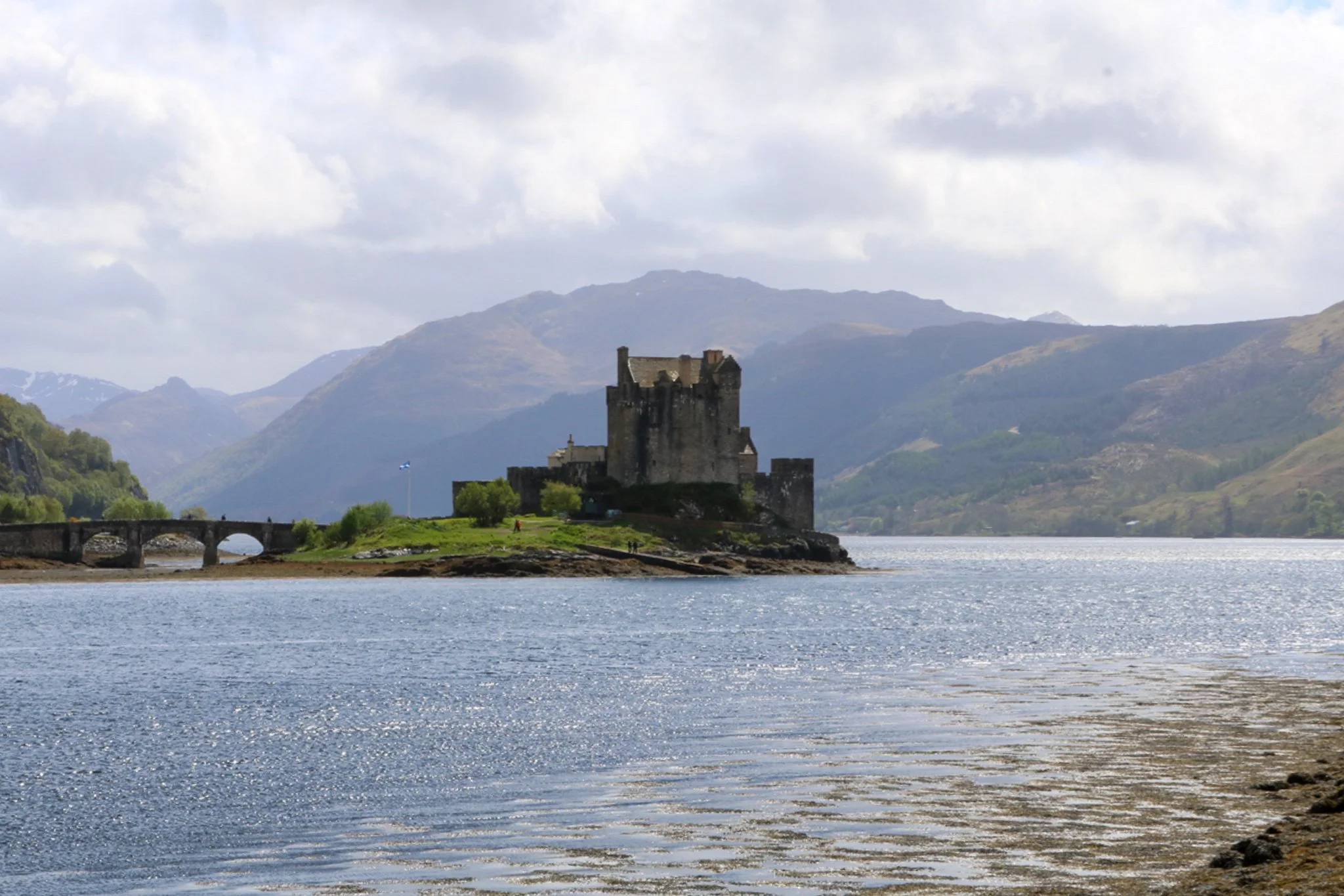 A castle on a small island in a body of water, with mountains in the background and a cloudy sky overhead.