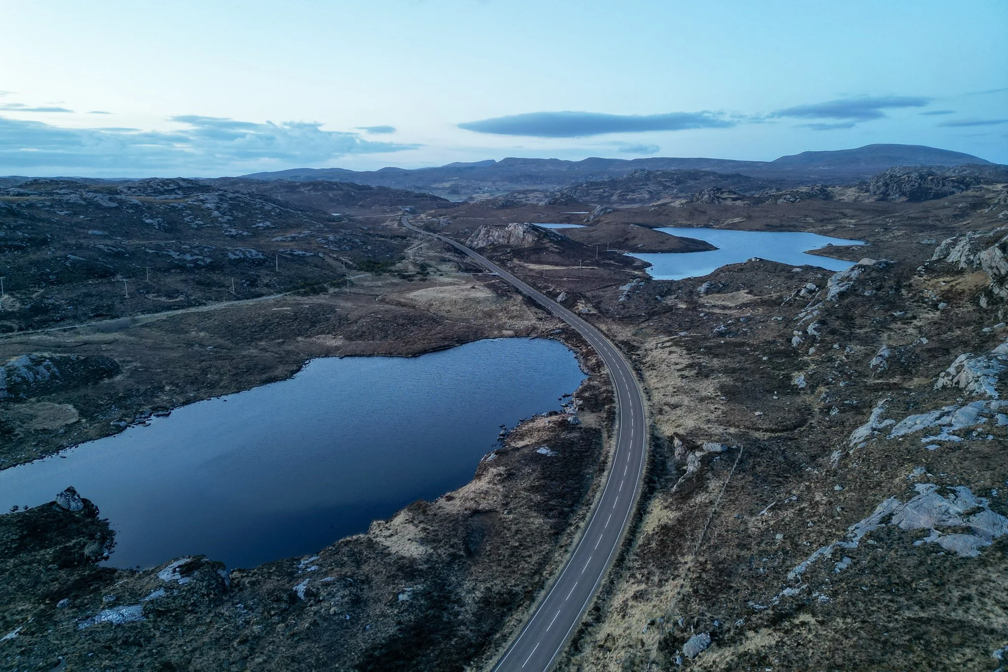 An aerial view of a winding road through a rugged landscape with lakes and hills under a cloudy sky.