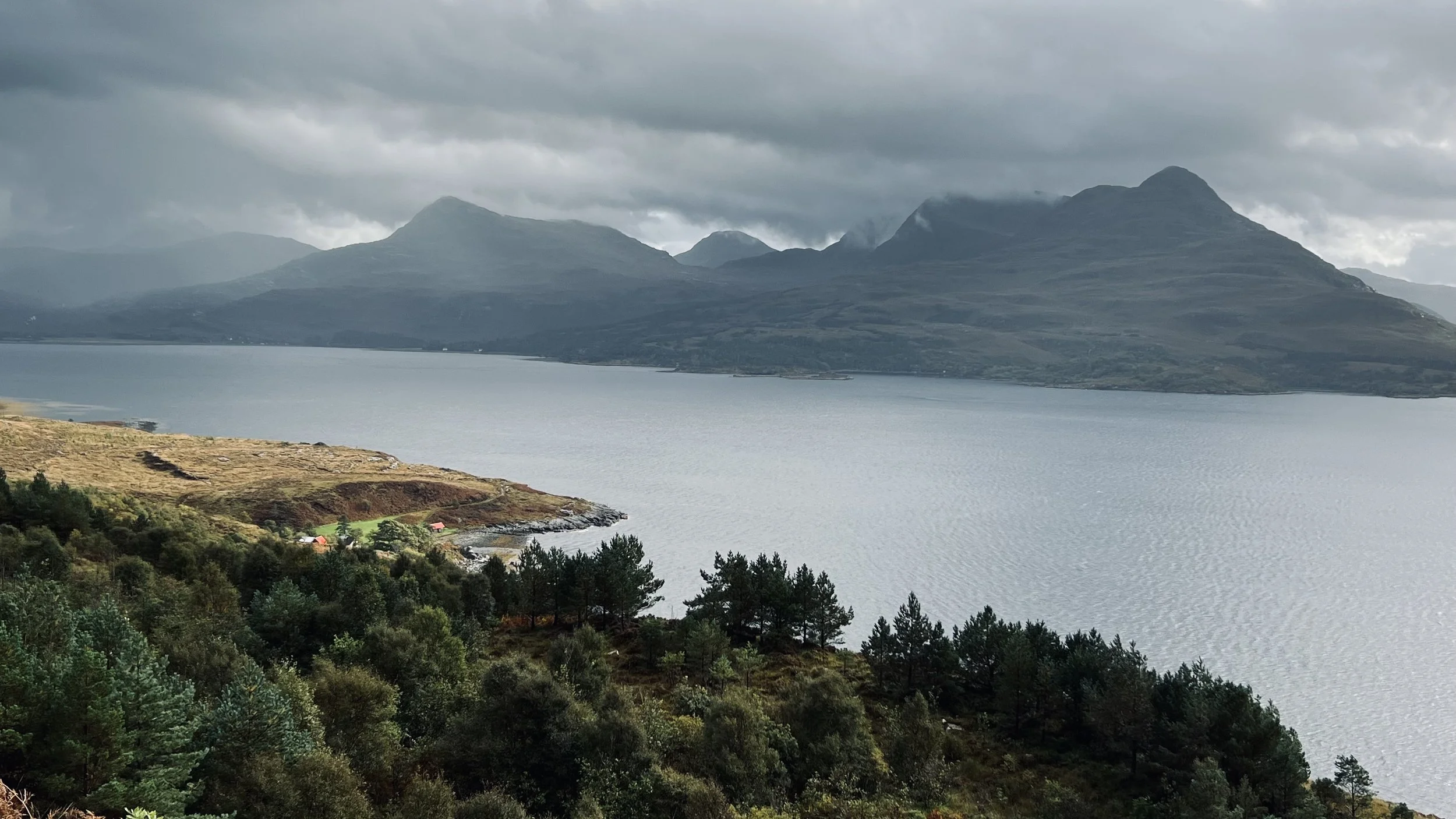 A scenic landscape with the epic mountains of Torridon in the background, a large body of water in the middle, and green forested hills in the foreground. The sky is cloudy and overcast.