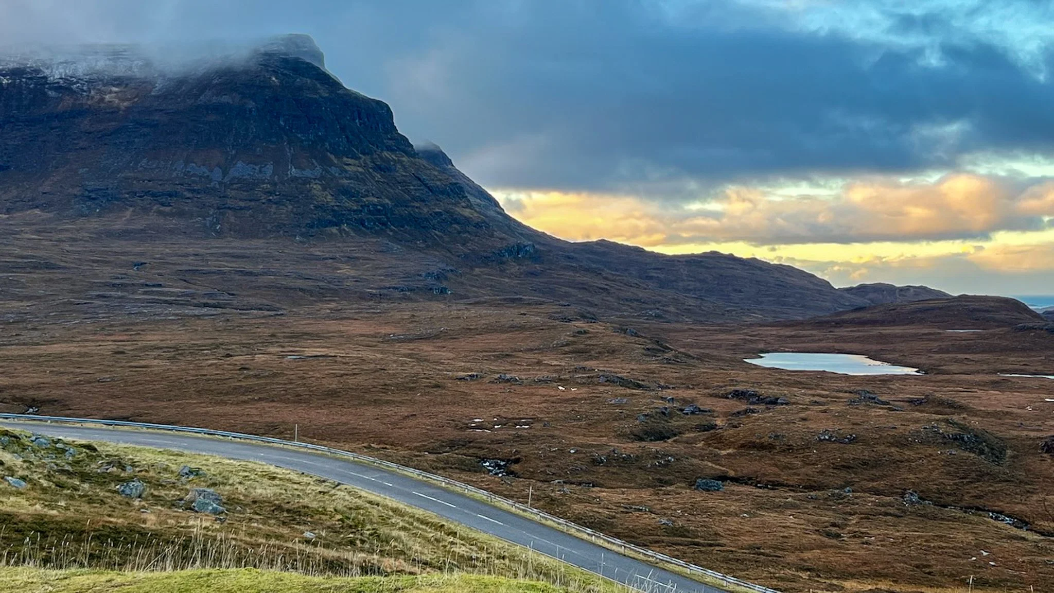 Scenic landscape featuring a winding road, open moorlands, mountains with partially snow-capped peaks, a small lake, and a partly cloudy sky during sunset.