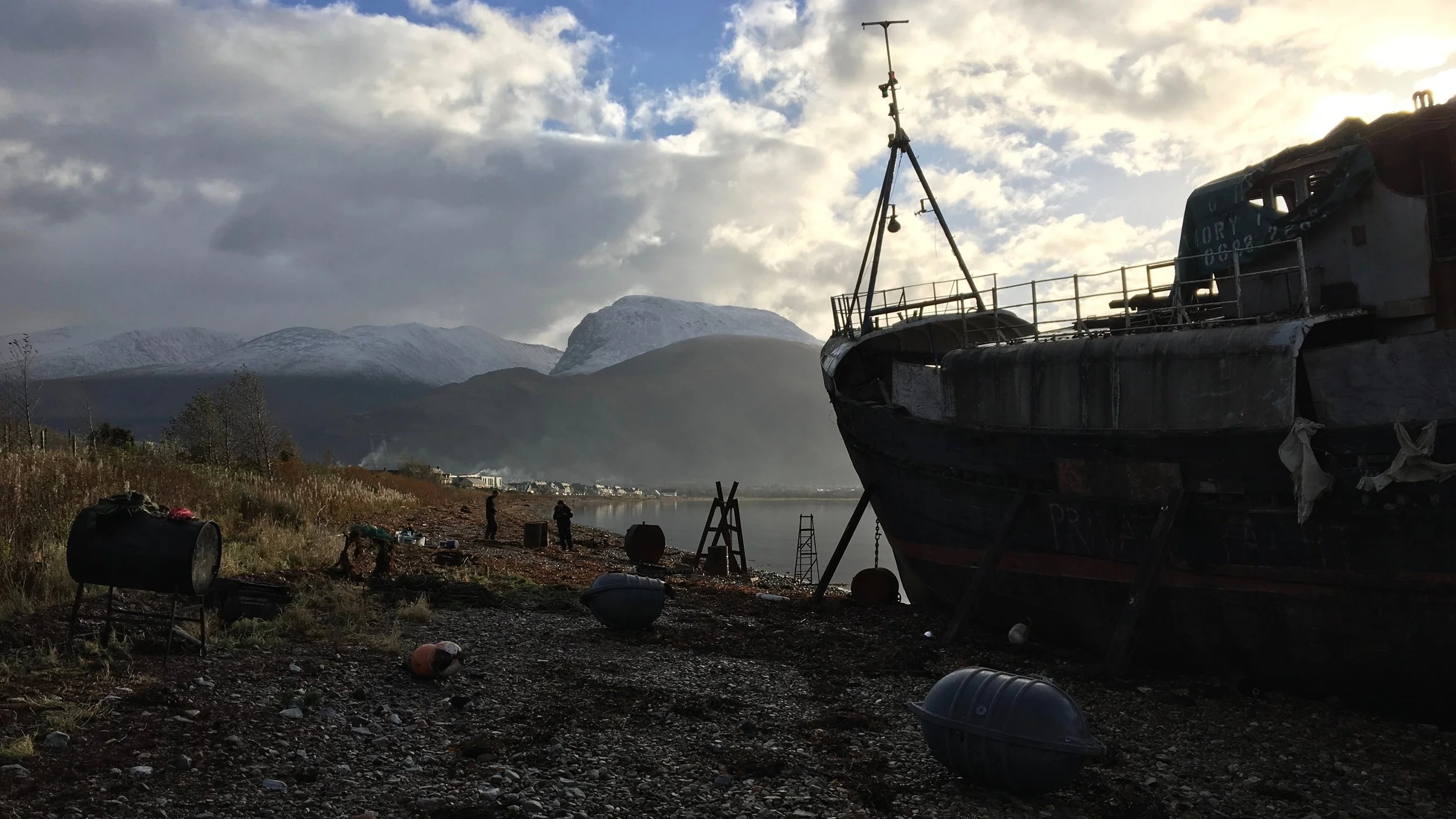 A large, weathered boat beached on a rugged shoreline with scattered debris including oil drums, against a backdrop of a cloudy skysnow-capped mountains including Ben Nevis .
