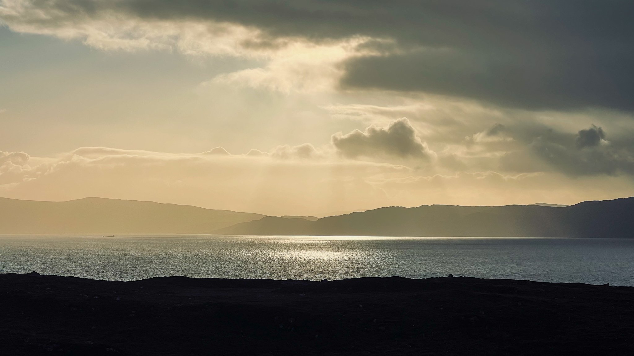 A scenic view of a body of water with hills in the background, under a cloudy sky with sunlight breaking through.