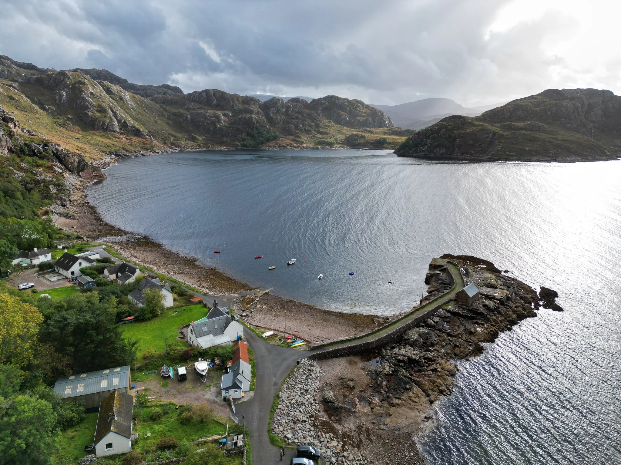 Aerial view of a small coastal village with houses, a rocky pier, and boats on a lake, surrounded by mountains and cloudy sky.
