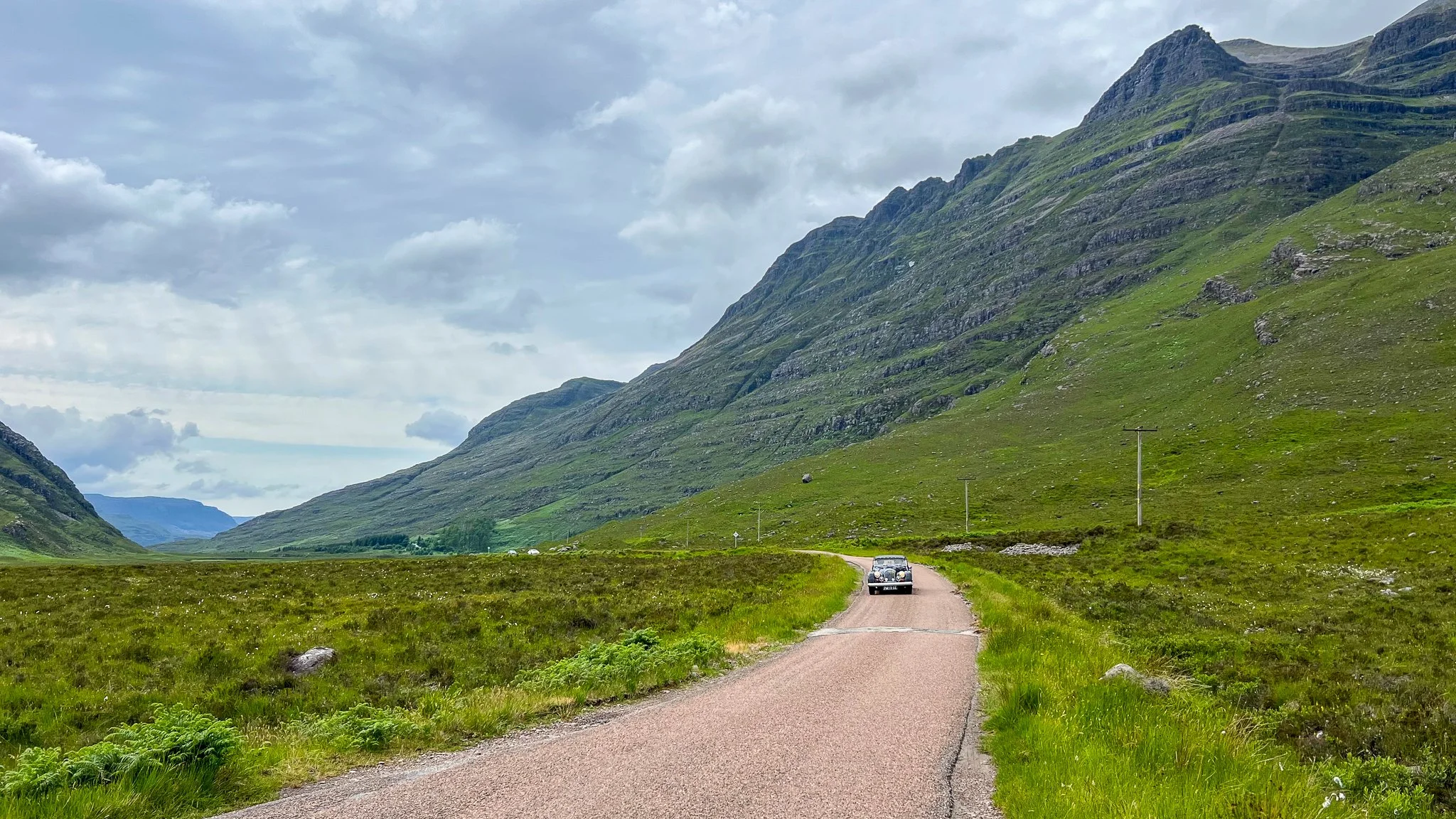 A narrow, winding road running through a green valley with mountains on both sides and a cloudy sky overhead. A small black car is driving on the road.