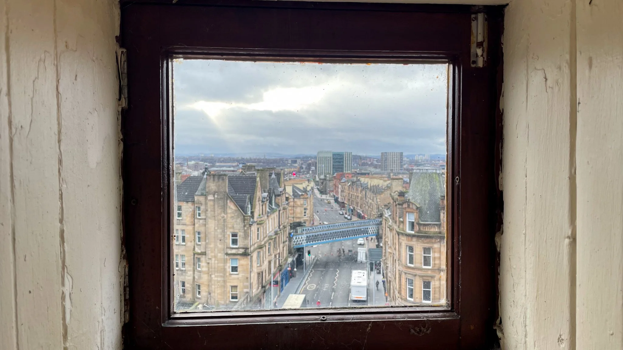 View of a city street through a small window with wooden frame and beige walls inside a building.