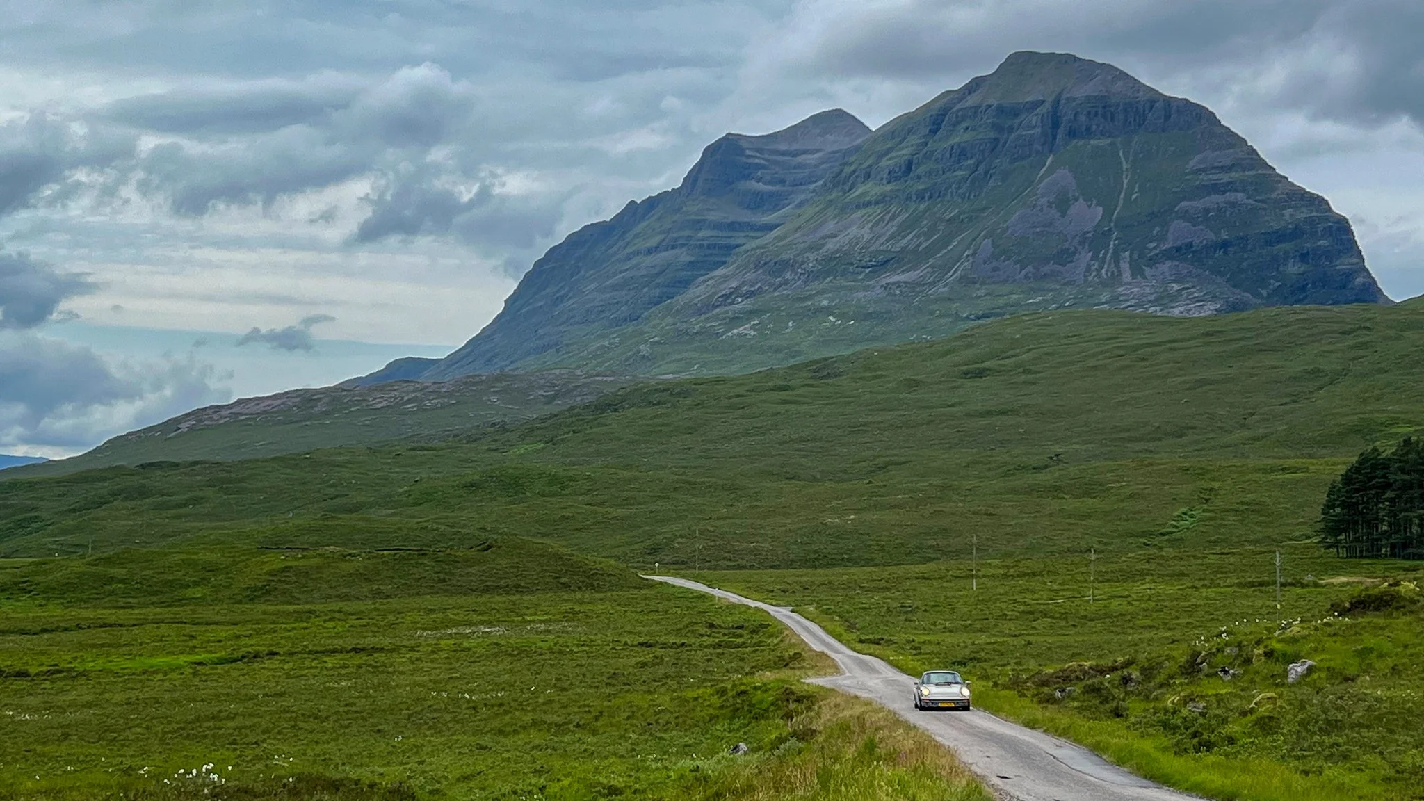 A car driving on a narrow, winding country road through green fields with a large mountain in the background under cloudy skies.