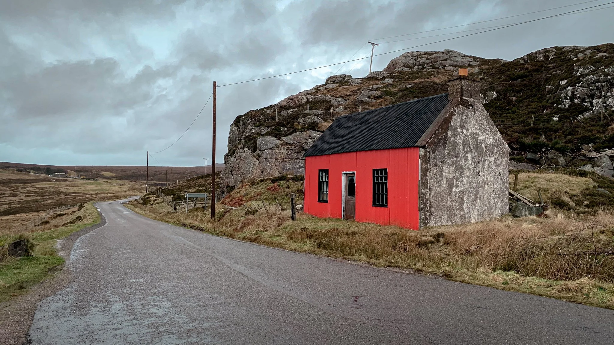 A small house with a red front and gray stone sides situated beside a winding country road in a rural landscape with hills and cloudy sky.