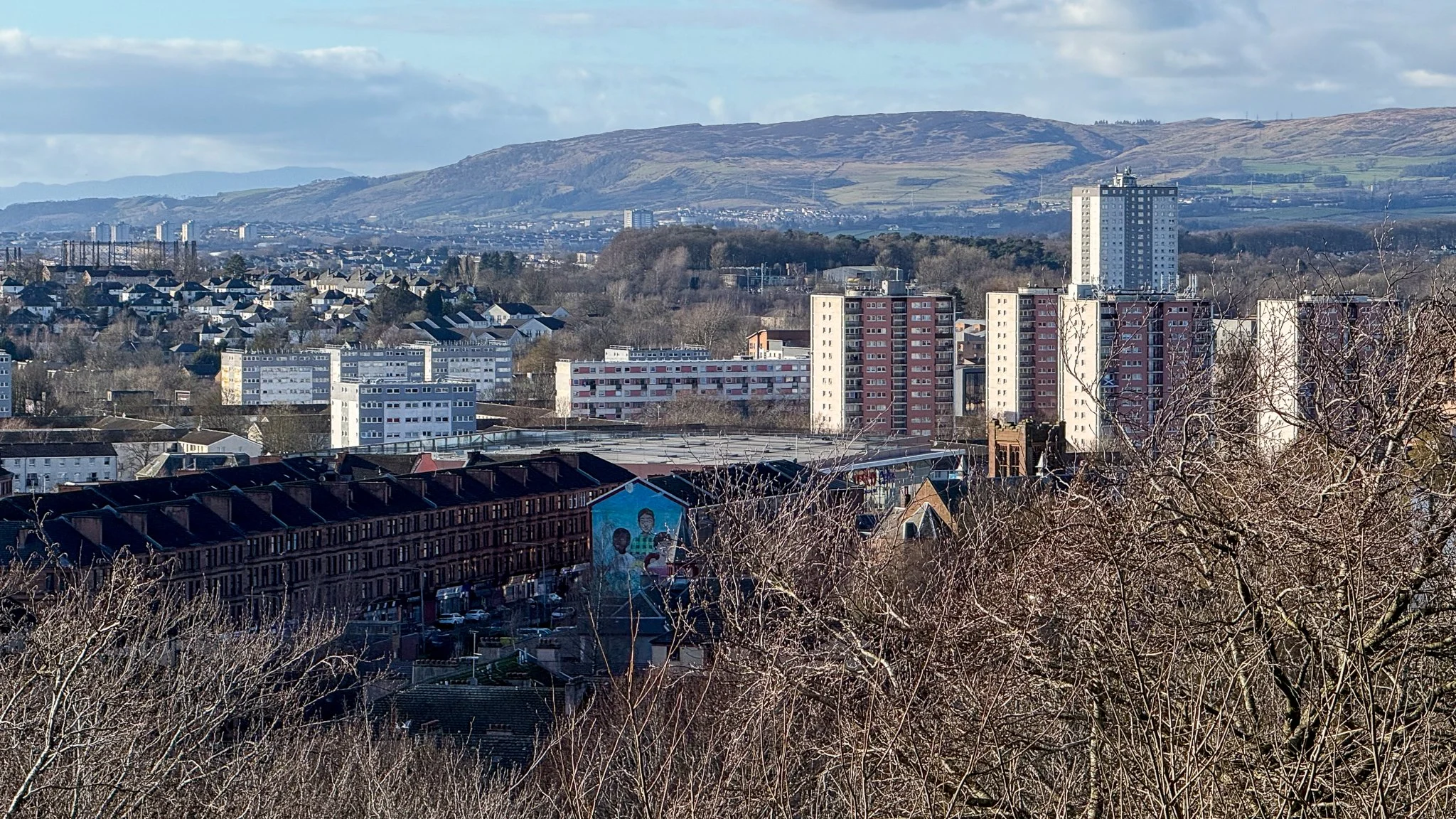 Cityscape with high-rise buildings and residential homes framed by leafless trees, hills in the background, and a partly cloudy sky.