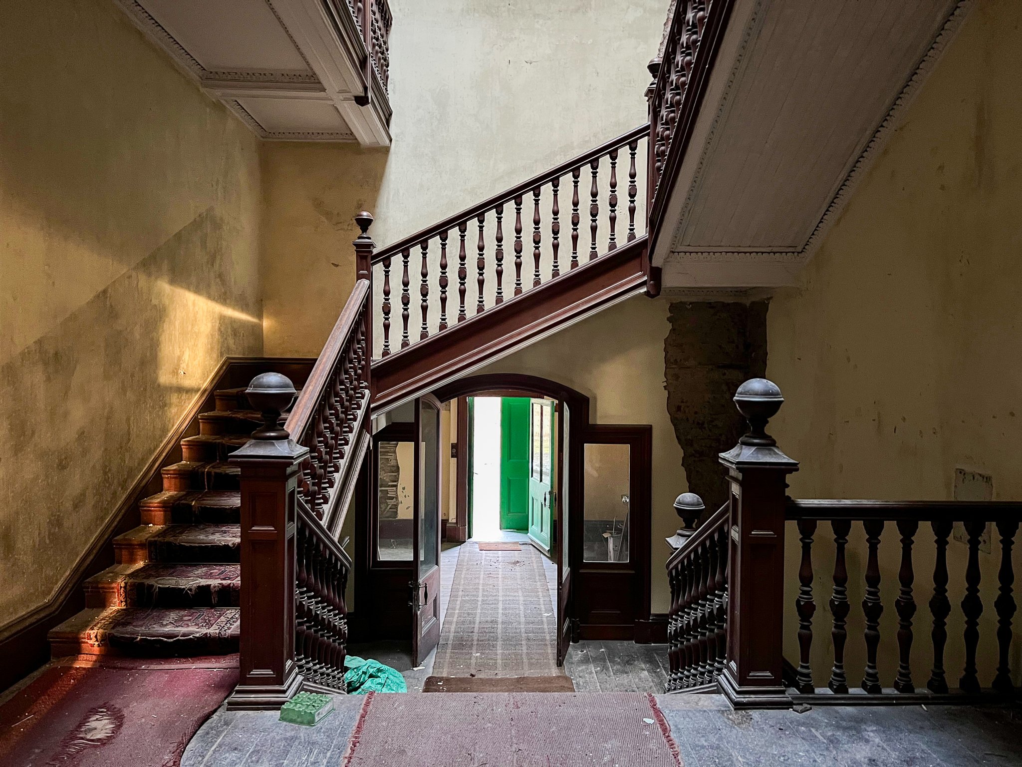 Interior of an old house staircase with wood railings, stairs with worn carpet, open green front door visible at the end of the hallway, and walls with peeling paint.