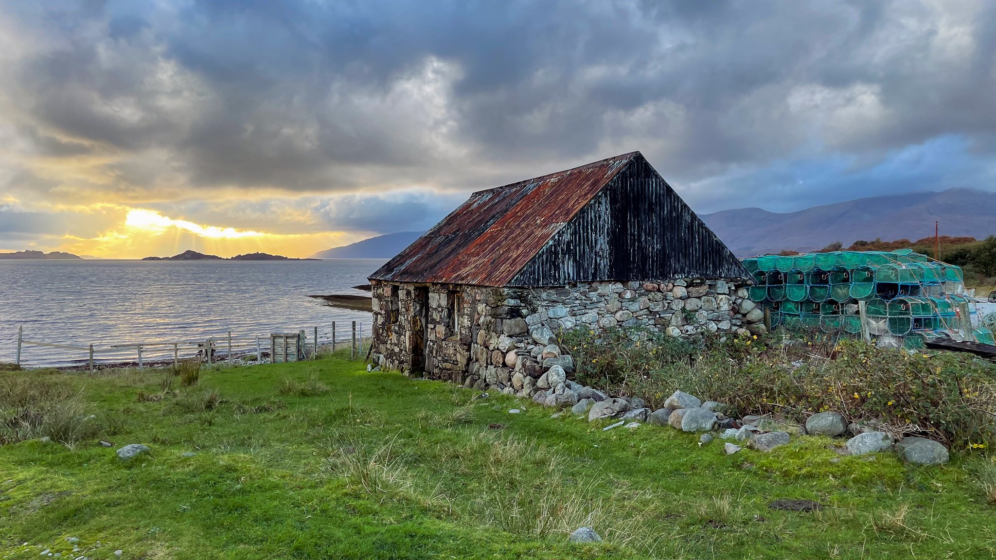 A stone and wood boathouse by the water with a sunset in the background, cloudy sky, and distant mountains.