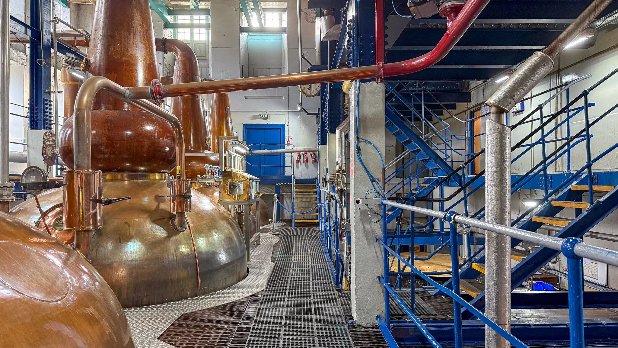 Interior view of a distillery with copper stills and blue metal staircases and railings.
