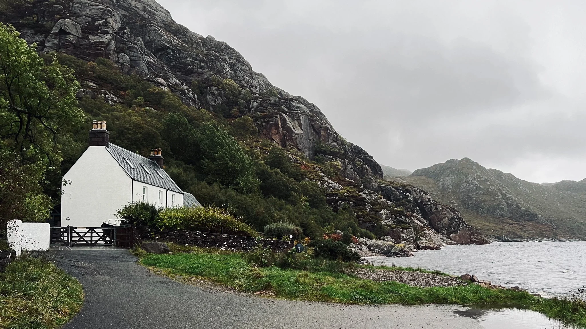 A white cottage with a gray roof and two chimneys, situated near water with a rocky hillside in the background on a cloudy day.