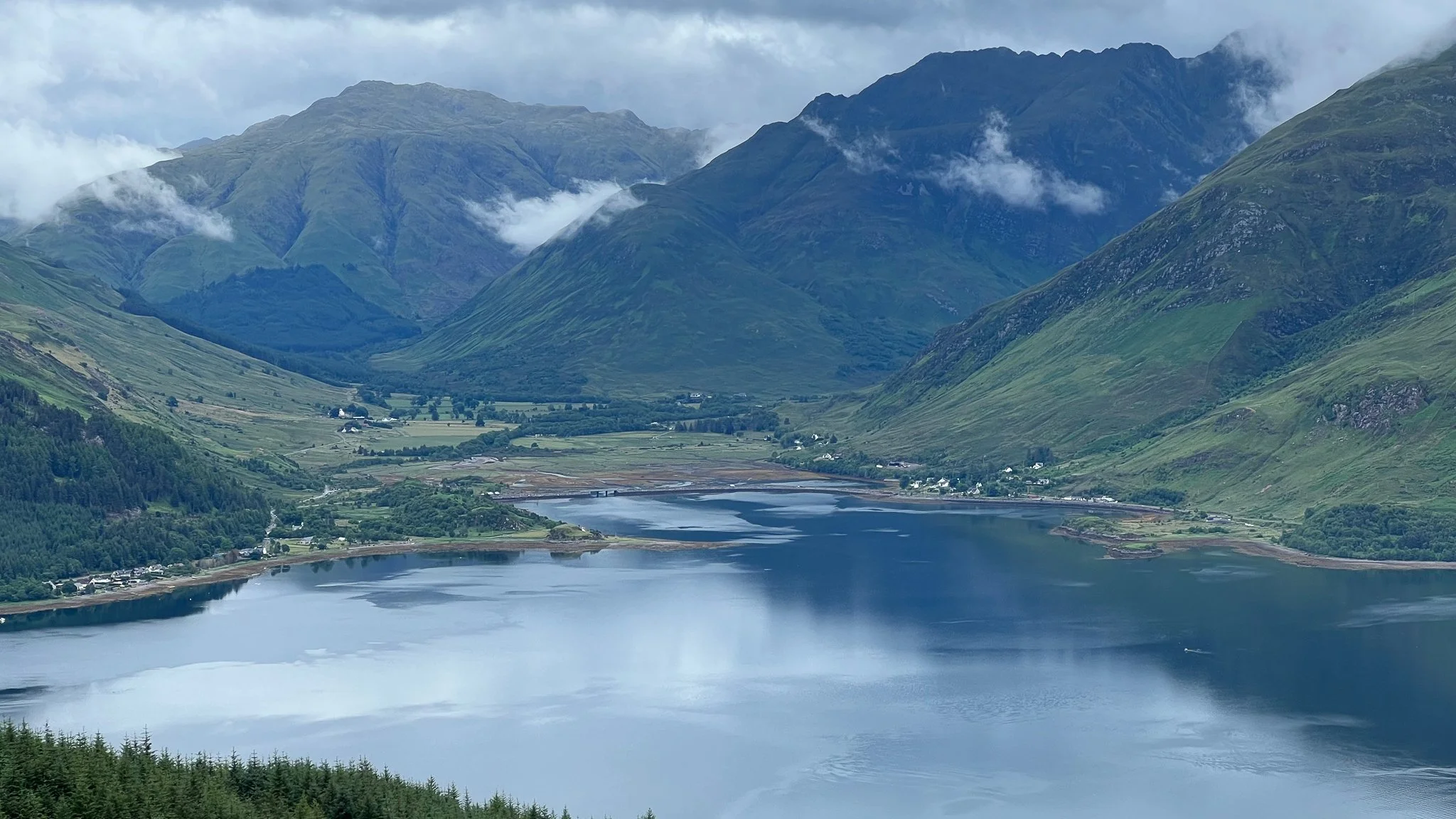 Scenic view of a lake in a valley surrounded by green mountains with cloudy sky.