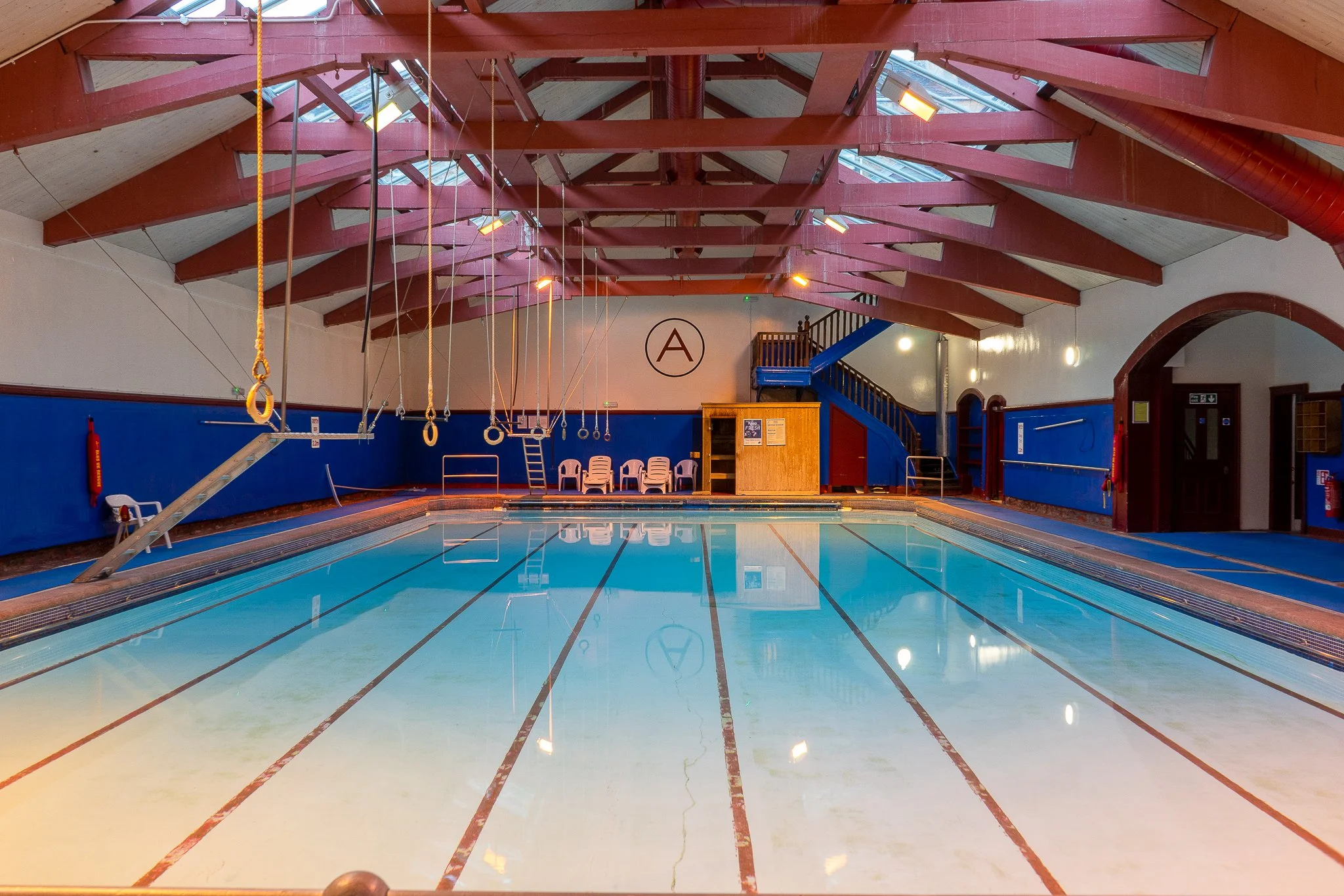 Indoor swimming pool with ropes and rings hanging from the ceiling, blue walls, red wooden beams, and a staircase in the background.