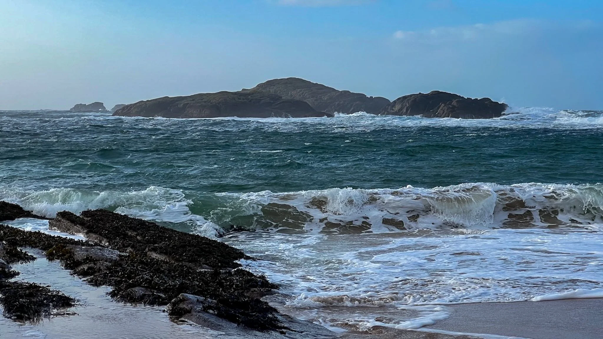 Waves crashing on a sandy beach with dark rocks, with an island or large rock formation in the distance under a blue sky.