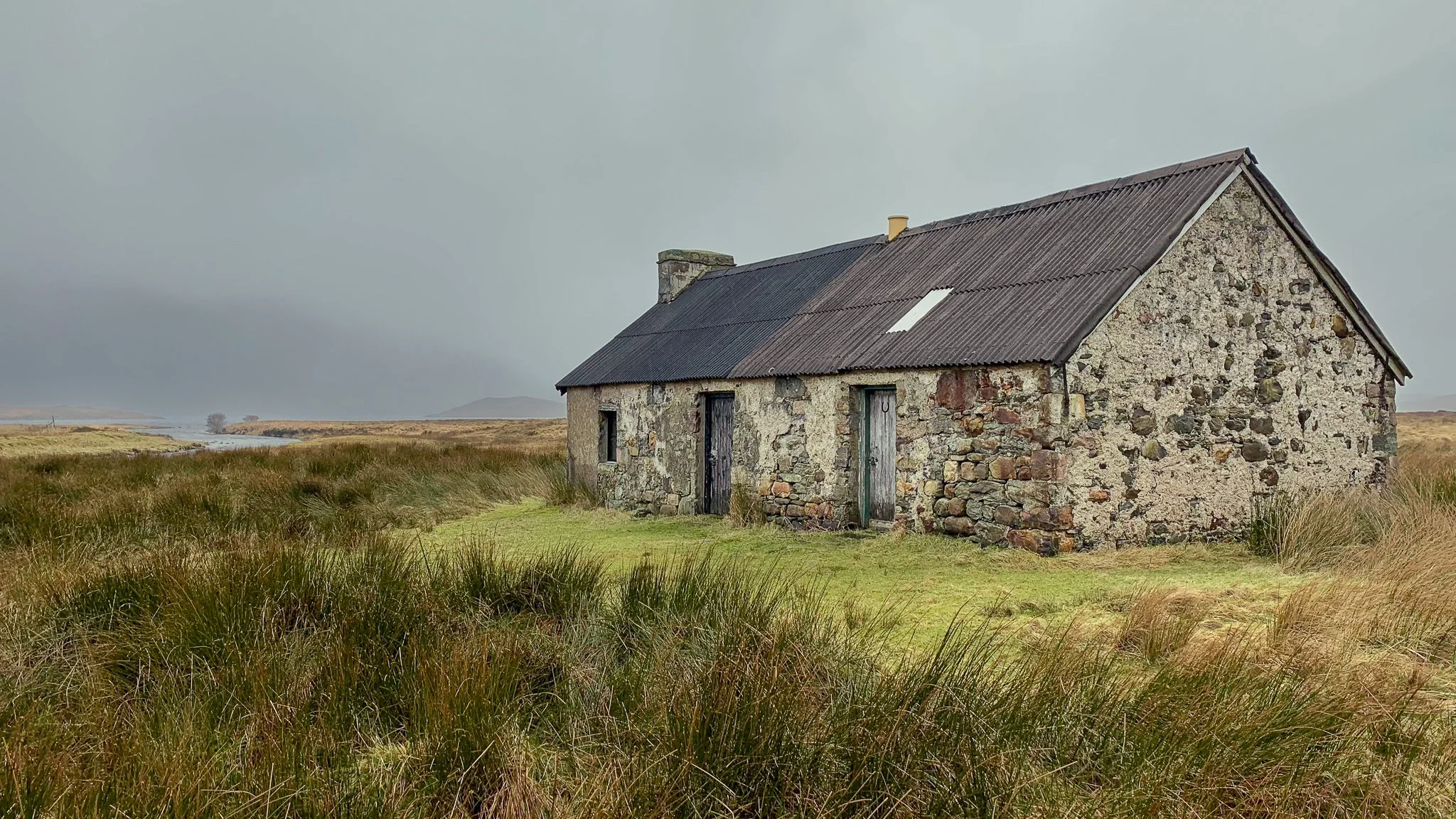 An old stone house with a corrugated metal roof situated in an open grassy landscape under a cloudy sky.