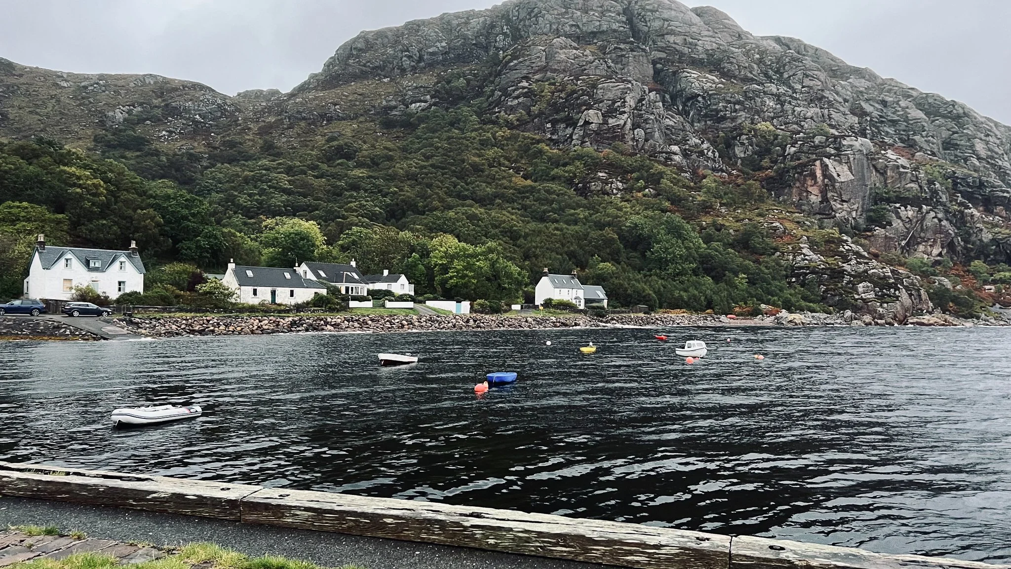 Scenic view of a small waterfront village with white houses, parked cars, and a rocky shoreline across a body of water with small boats, against a backdrop of steep, green mountains.