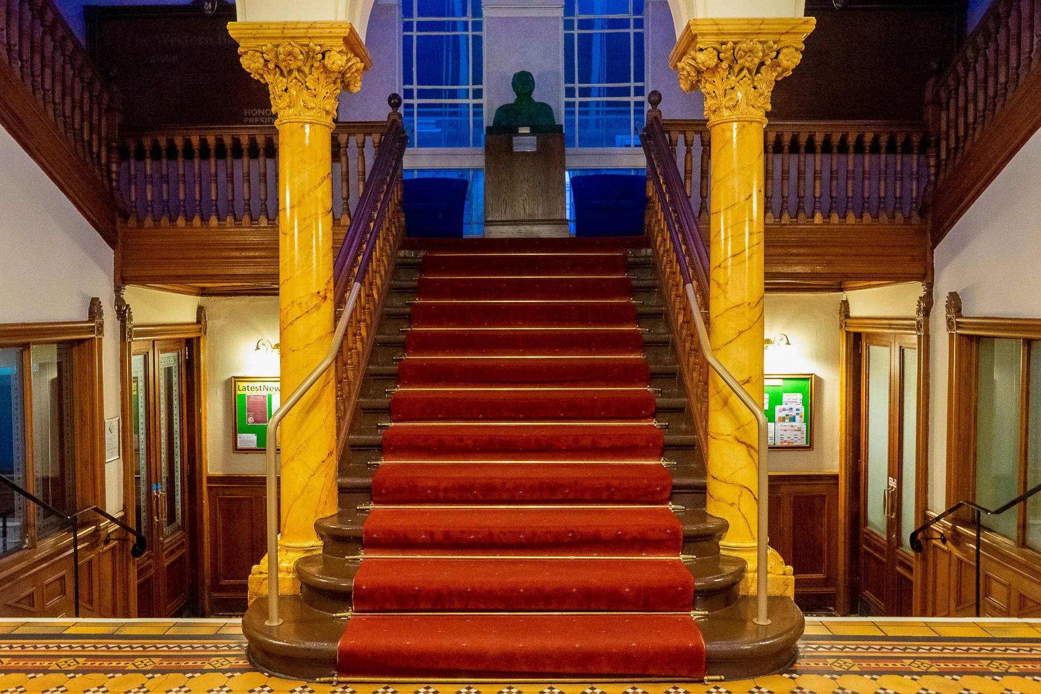 Interior view of a grand staircase with red carpet, yellow marble columns, wooden railings, and glass windows at the top, reflecting an elegant and historic building.