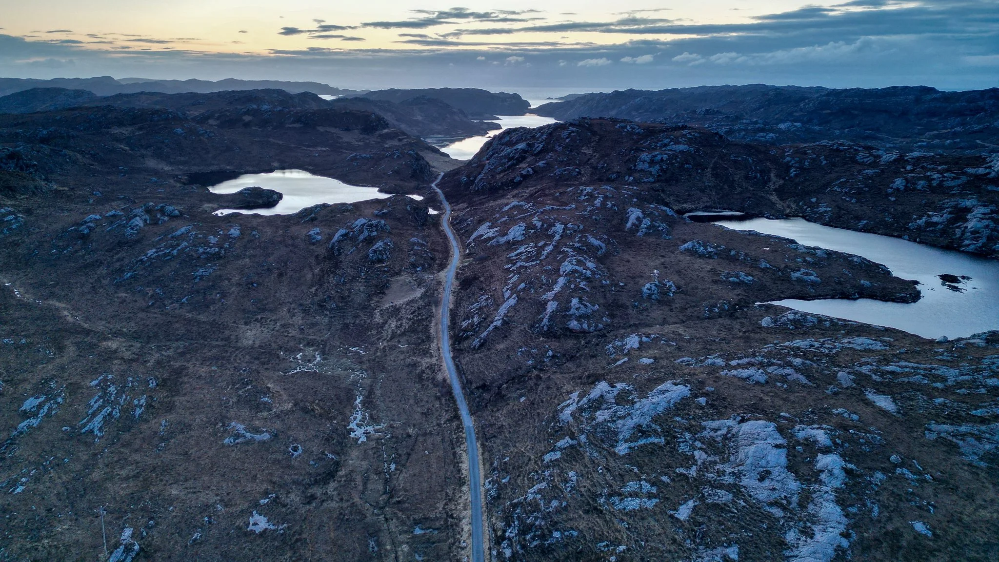Aerial view of rugged mountain landscape with lakes and a winding road at dusk.