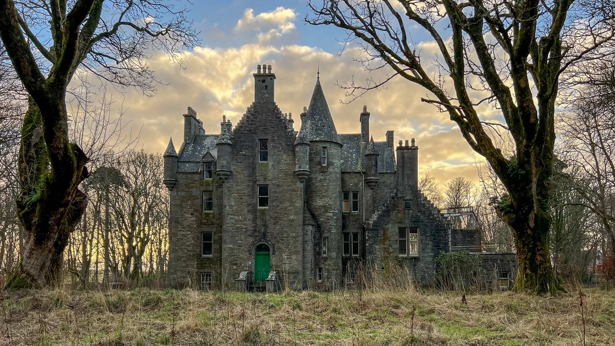 A large, dark stone castle with tall, pointed turrets situated in a wooded area. The sky features clouds and a setting or rising sun, creating a silhouette effect. The castle has a green door and appears abandoned.