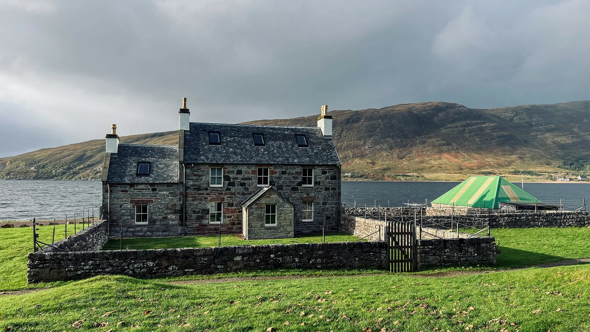A stone house near a body of water with hilly terrain in the background. The house has a slate roof and multiple small chimneys. There is a fenced grassy yard with a wooden gate, and a green and yellow tent-like structure nearby.