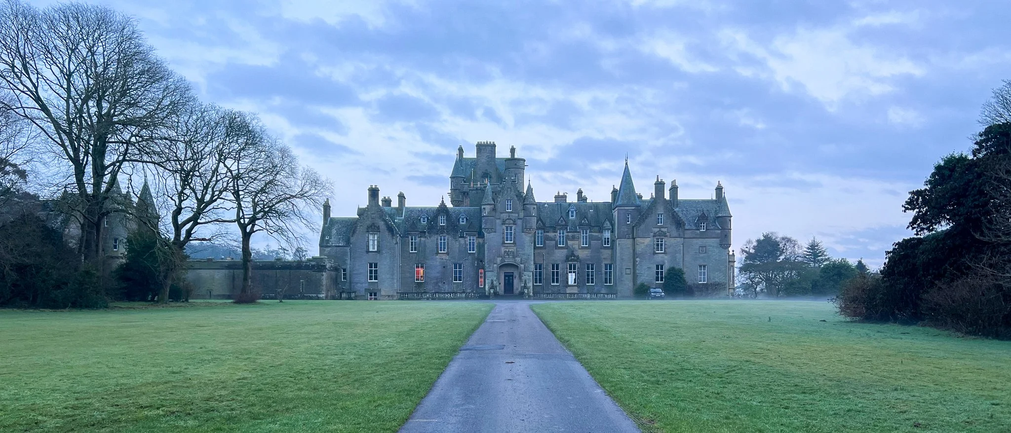 A large, historic castle with pointed towers and multiple windows, set in a green landscape with a pathway leading up to it. The sky overhead is partly cloudy, and there are leafless trees on either side of the castle.