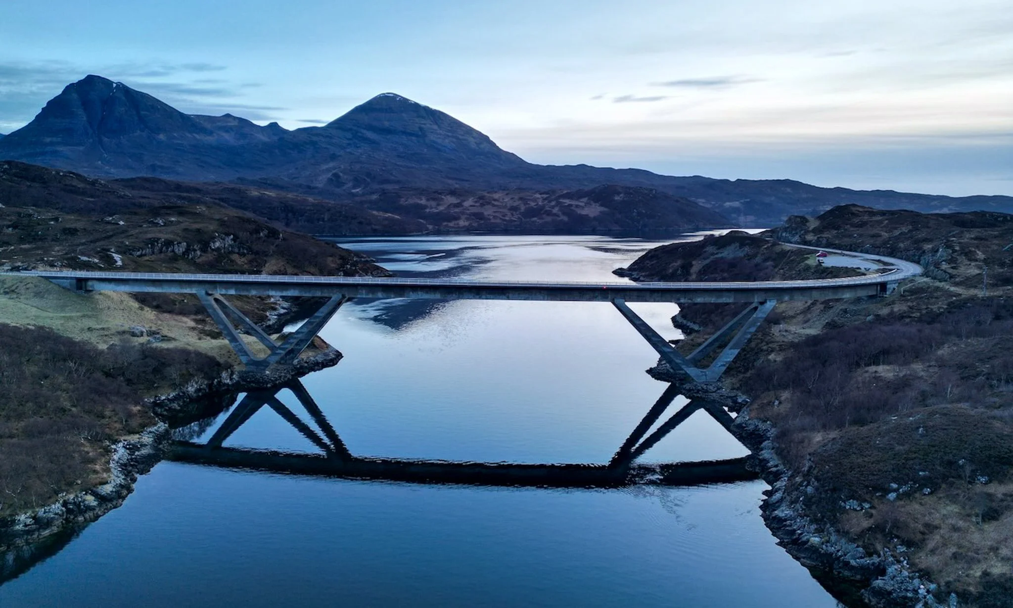 Kylesku bridge arching over a body of water with mountains in the background, taken during twilight.