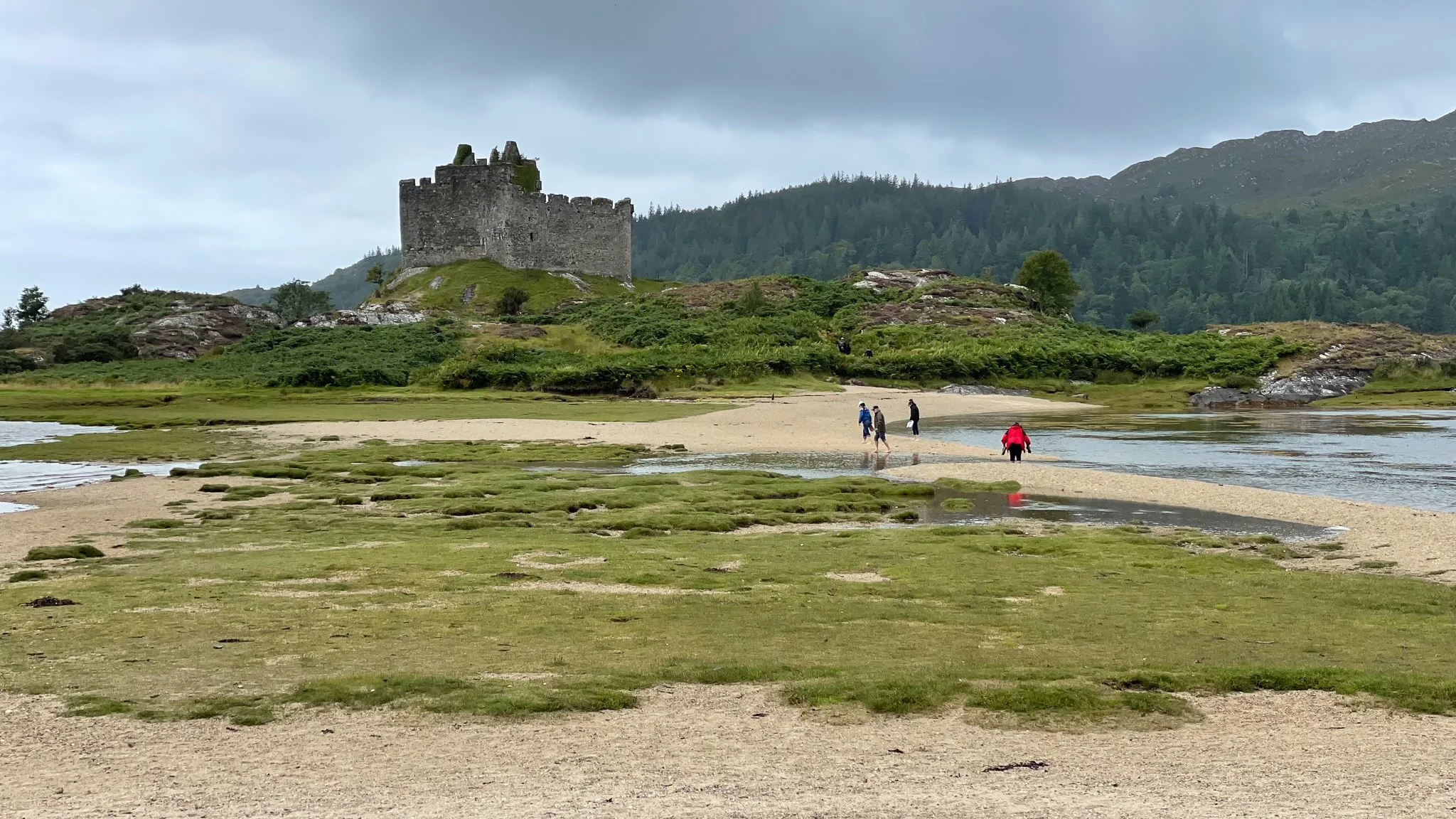 A medieval castle on a hill overlooking a sandy and grassy area with small pools of water, with a river and forested mountains in the background and several people walking along the riverbank.