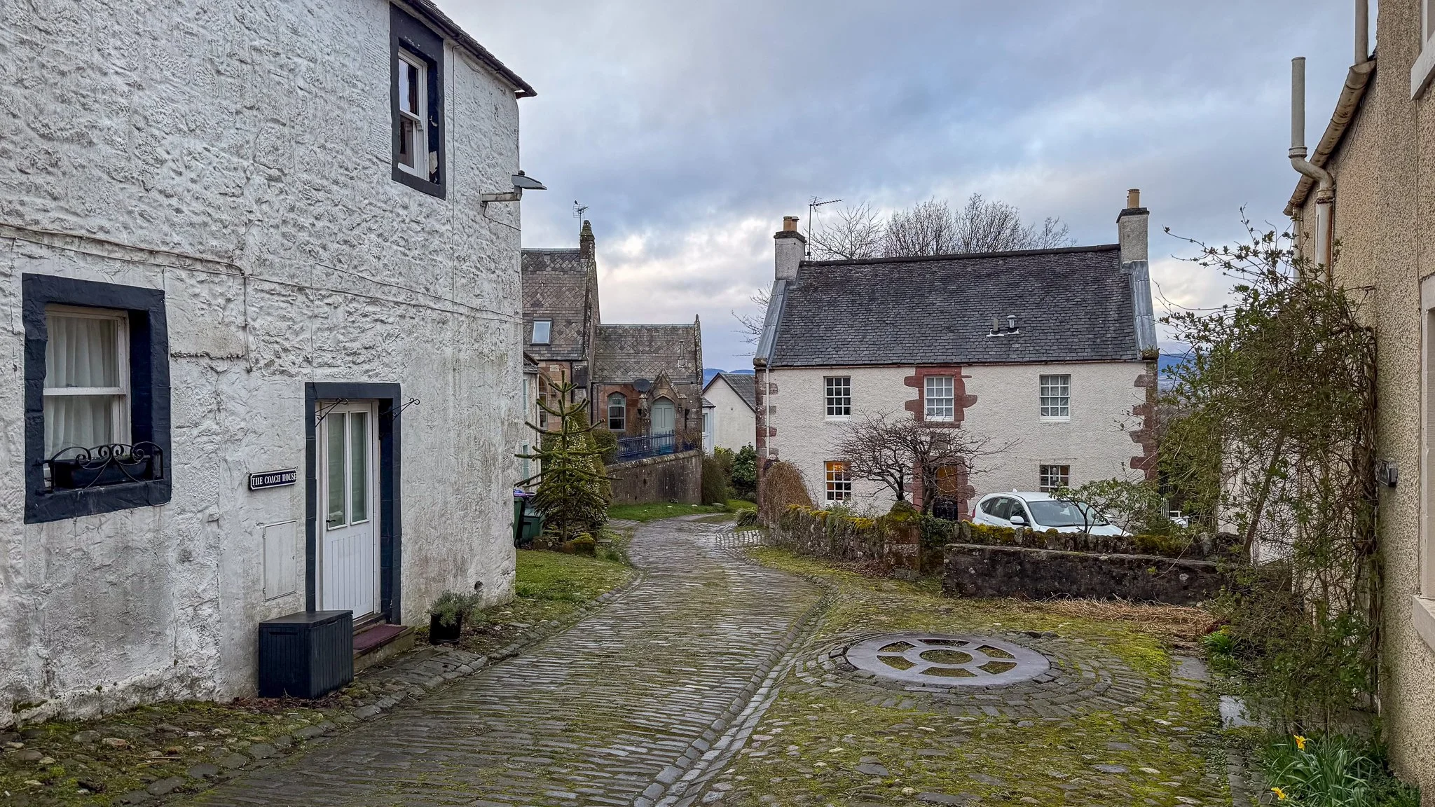 A cobblestone alleyway with old houses on either side, one with a sign reading 'The Coach House'. There are trees, bushes, and a parked car visible in the background, under a cloudy sky.