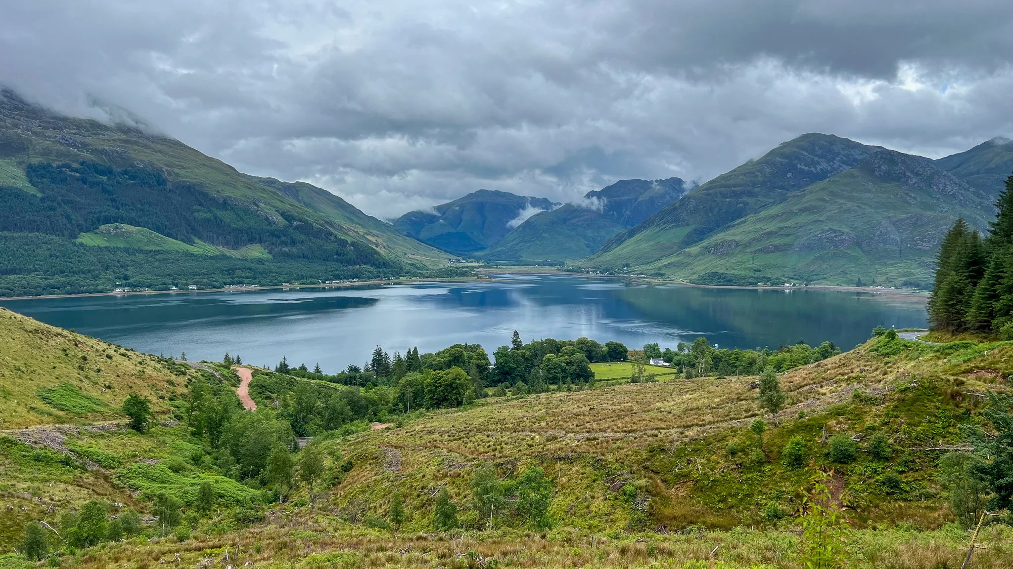 A scenic view of a lake surrounded by green hills and mountains under a cloudy sky, with a winding dirt path visible in the foreground.