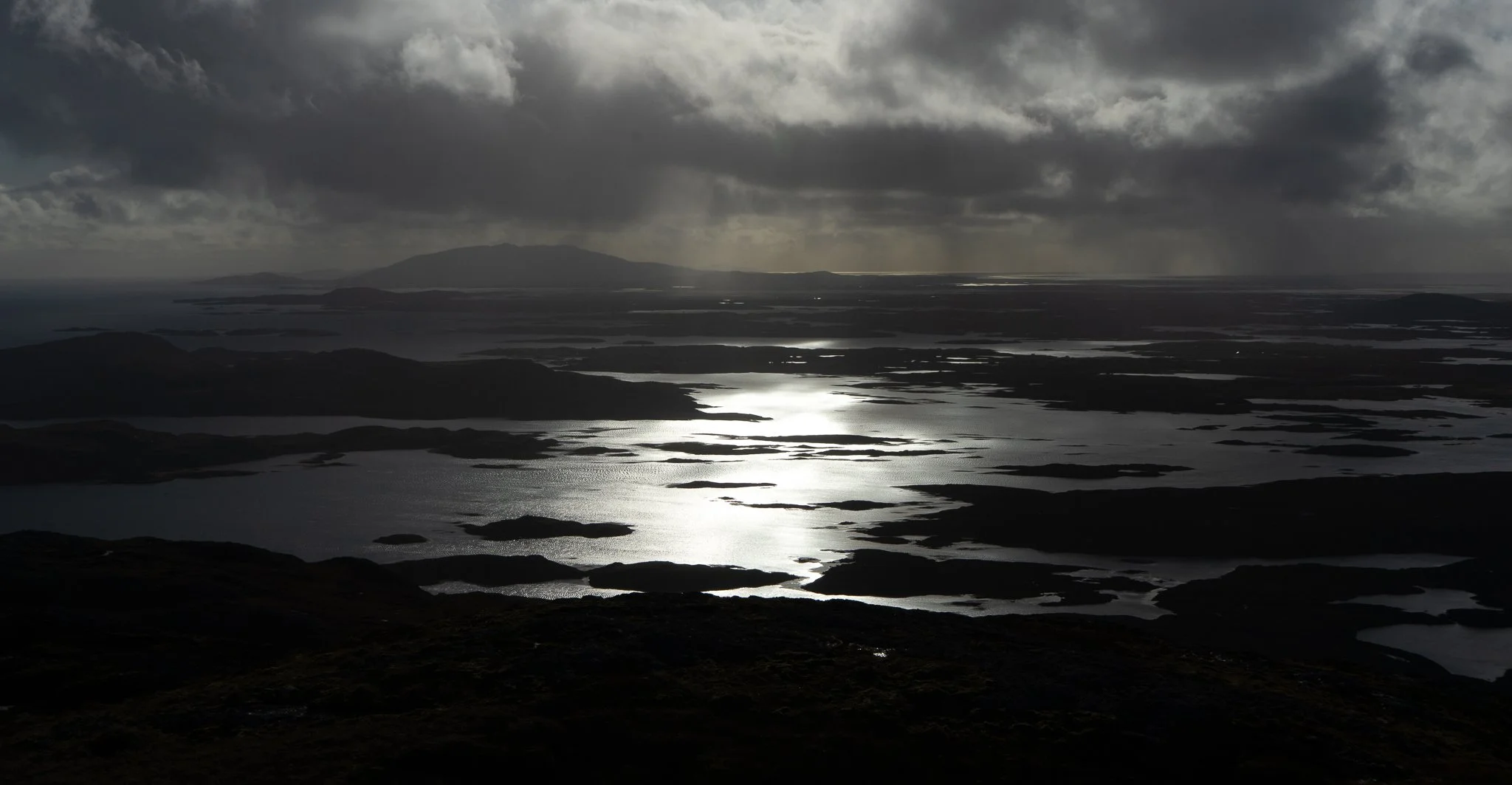 A landscape view of a dark, cloudy sky over a body of water with numerous islands and landmasses reflecting light from the overcast sky.