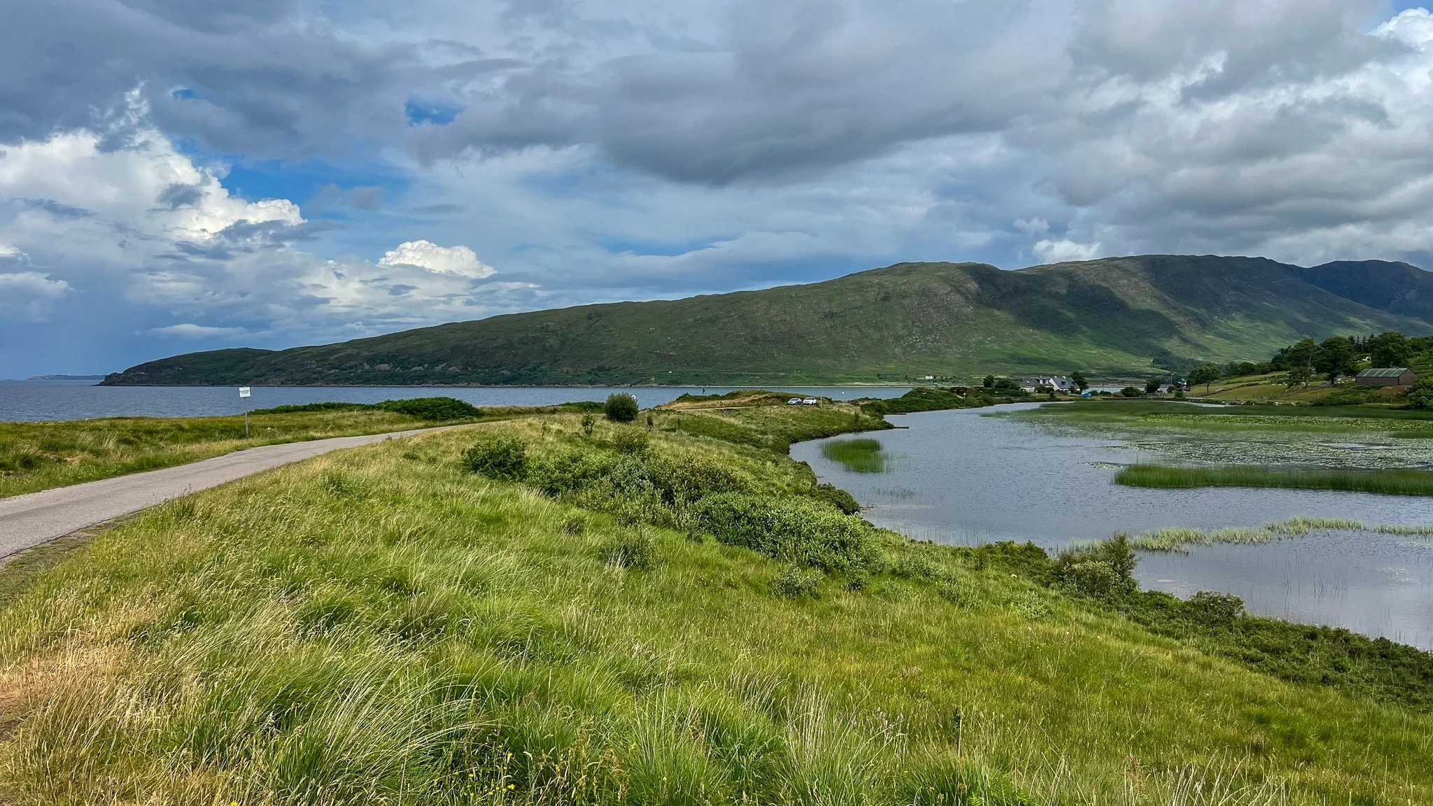 A scenic landscape featuring a grassy pathway beside a body of water with a mountain range in the background and partly cloudy sky.