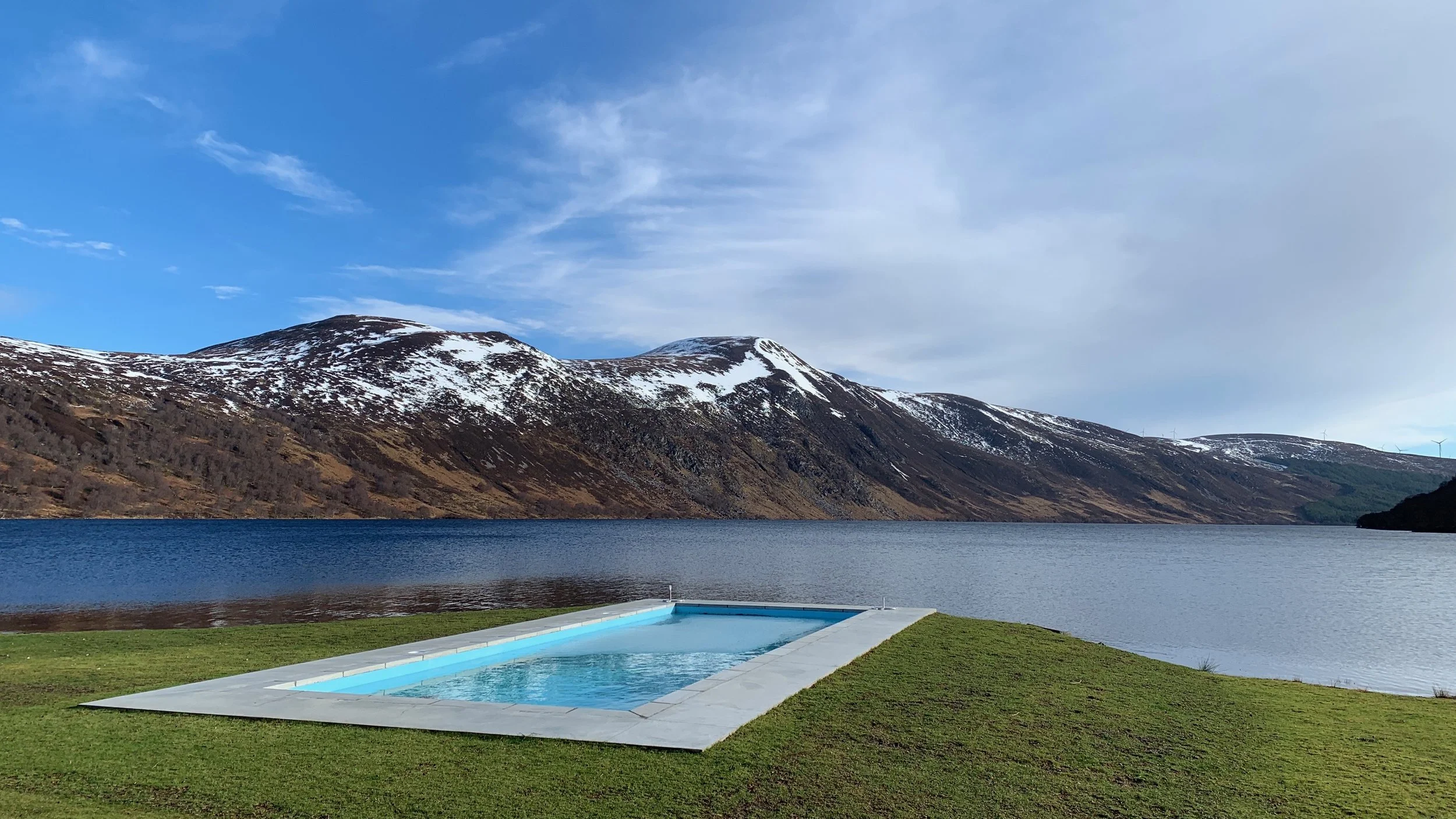 A rectangular swimming pool with clear blue water is situated on manicured green grass near a large lake or loch. In the background, snow-capped mountains and a partly cloudy sky are visible.